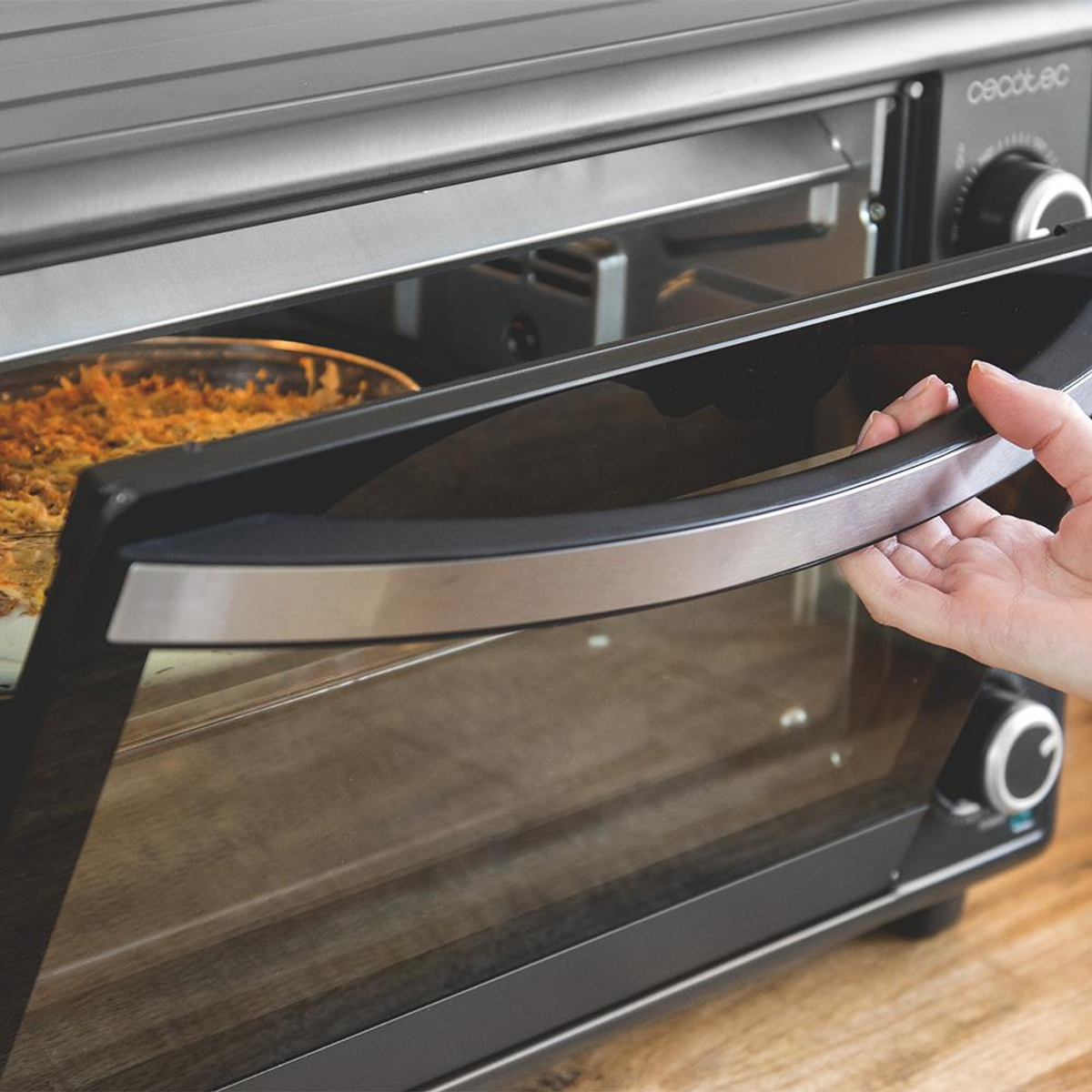 Ręka otwiera drzwi piekarnika, odsłaniając naczynie z jedzeniem. Piekarnik jest czarny ze srebrnym uchwytem. Tło to drewniana powierzchnia.