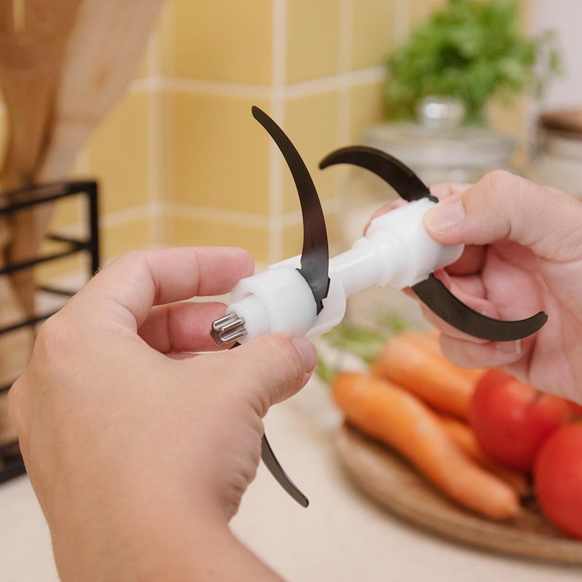 Manos sosteniendo una cuchilla blanca y negra de procesador de alimentos. Zanahorias y tomates en el fondo.