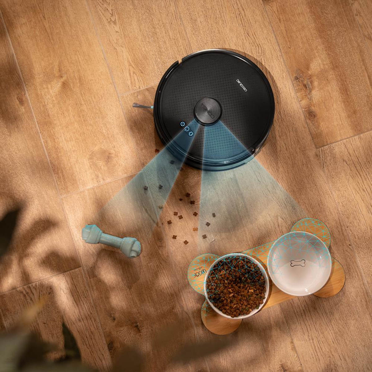 Un aspirapolvere robot, ciotole per cani e un giocattolo su un pavimento in legno. L'aspirapolvere emette raggi blu. Briciole di cibo sparse.