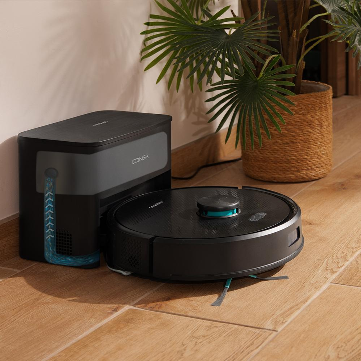 Un aspirapolvere robot nero accanto alla sua base di ricarica. Una pianta da interno in un vaso di vimini sullo sfondo su pavimento in legno.