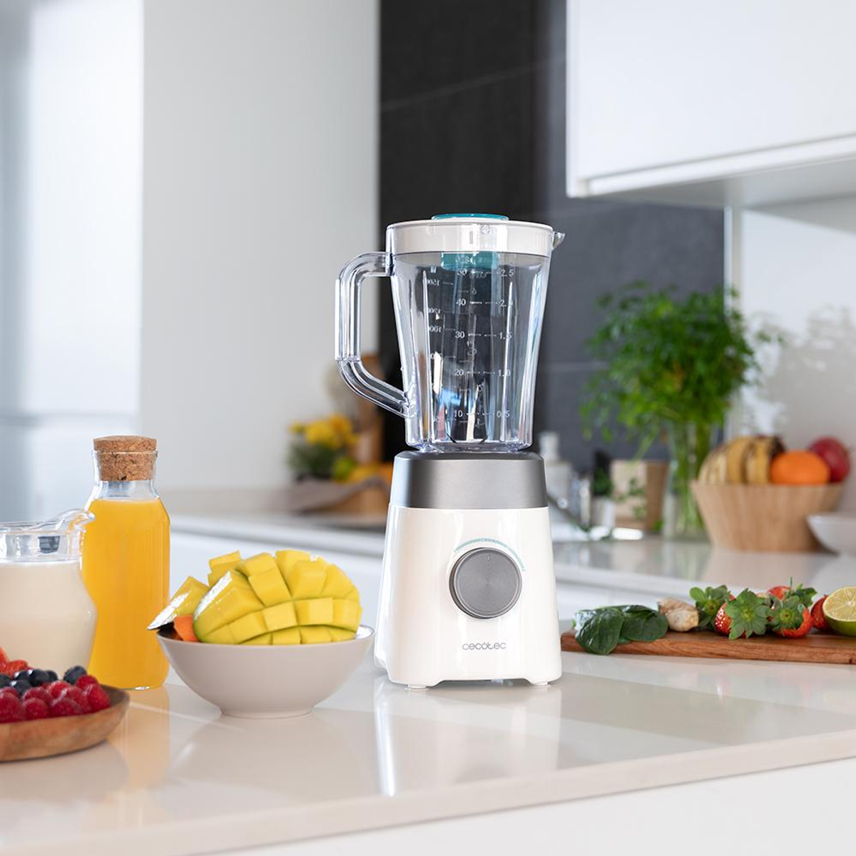 Un frullatore bianco con un barattolo di vetro è su un ripiano bianco. Mango a fette, frutta, succo e bacche sono disposti intorno.