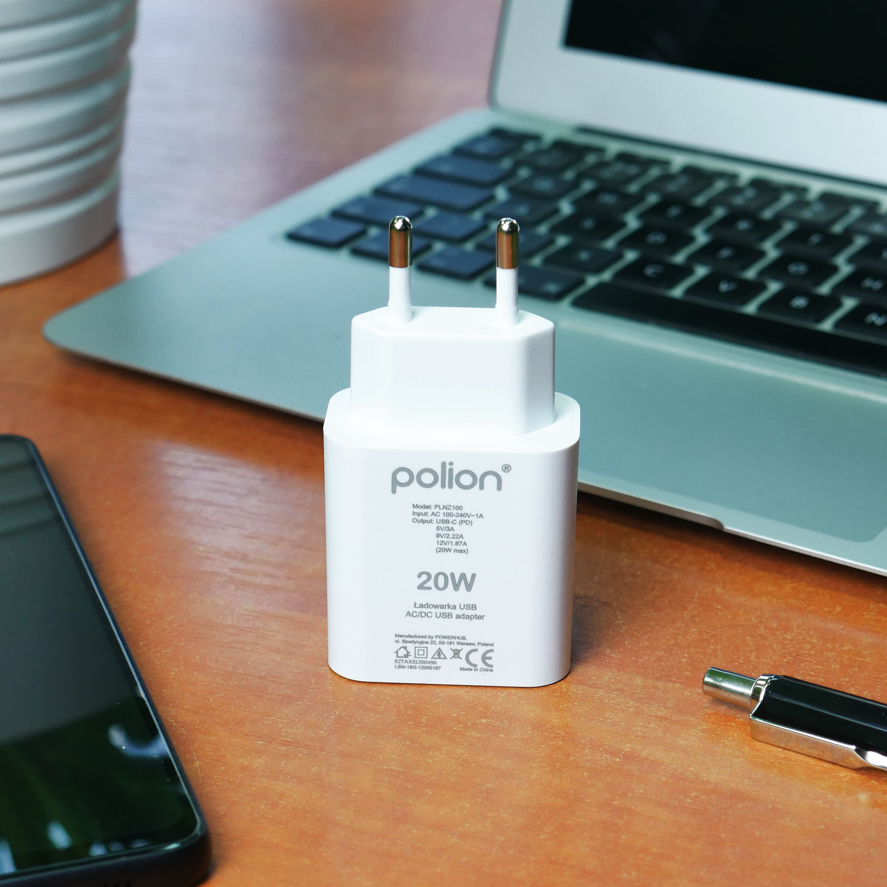 Biała ładowarka USB Polion z bolcami leży na drewnianym biurku obok laptopa i telefonu.