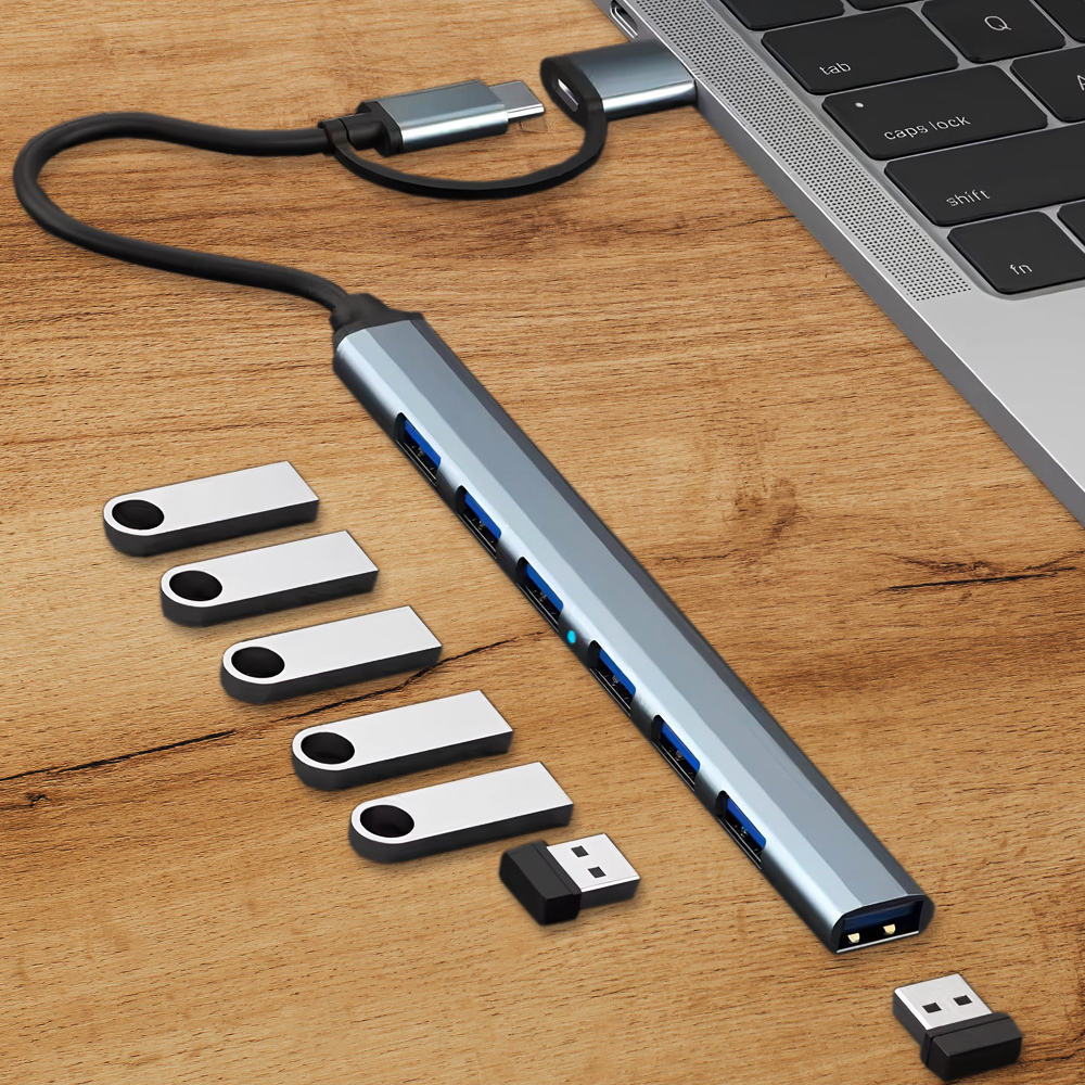 Srebrny hub USB podłączony do laptopa. Kilka srebrnych pendrive'ów i czarny adapter USB na drewnianej powierzchni.