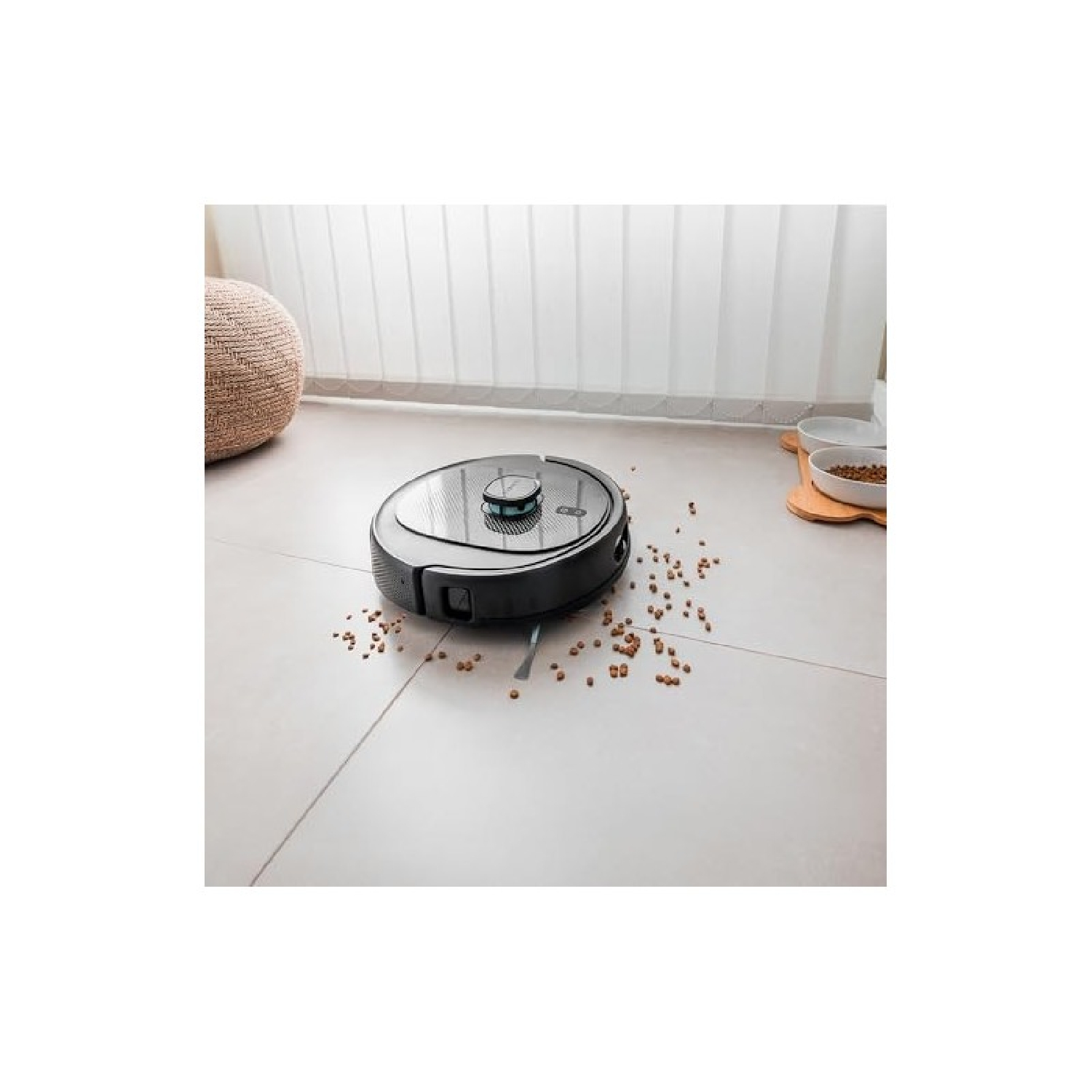 Un robot aspirador en un suelo embaldosado. Comida esparcida, un taburete tejido y cuencos para mascotas en el fondo.