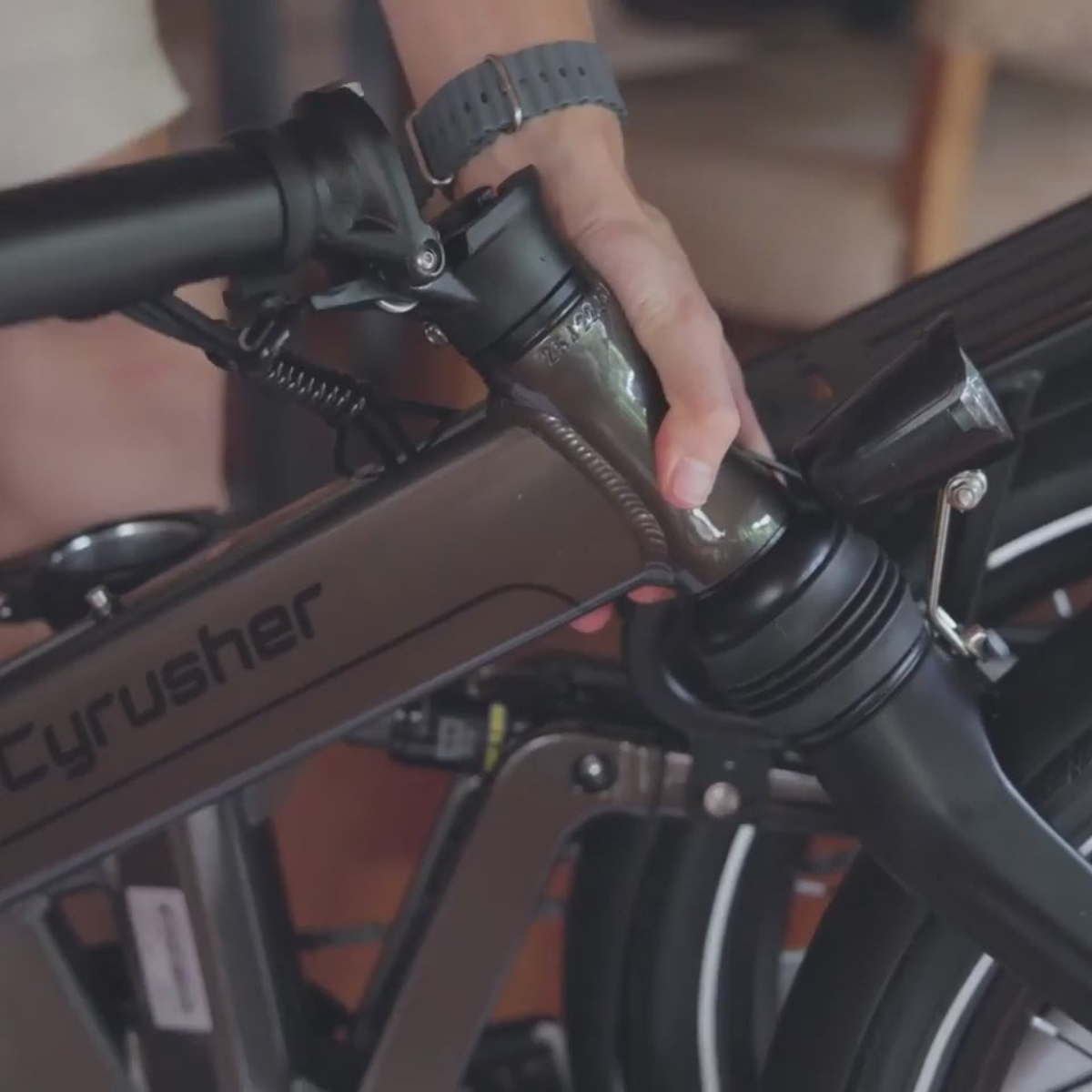 Ręka osoby reguluje kierownicę ciemnoszarego roweru składanego. Na rowerze widnieje nazwa "Cyrusher".