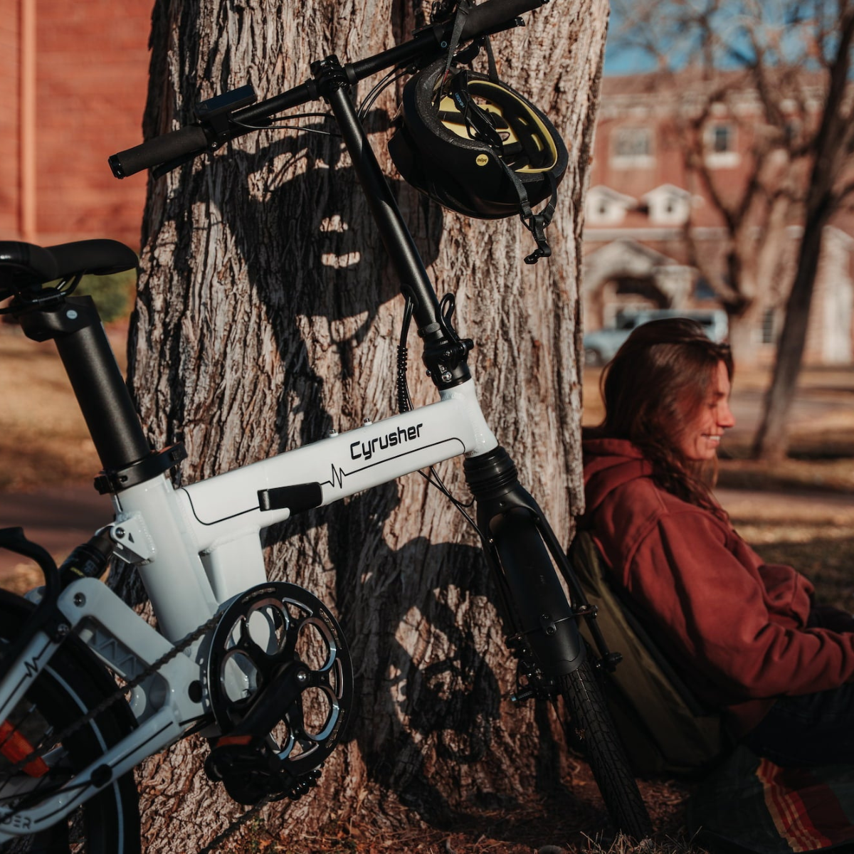 Biały rower opiera się o drzewo. W pobliżu siedzi osoba w czerwonej bluzie z kapturem, uśmiechając się. Kask wisi na rowerze.