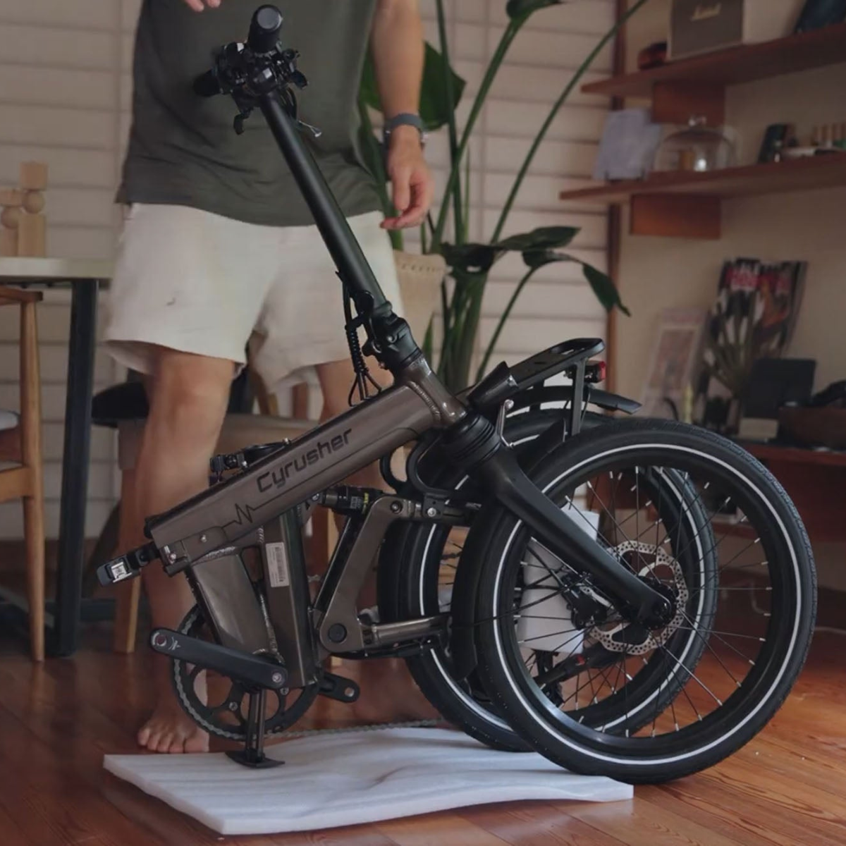 Mężczyzna trzyma złożony rower elektryczny Cyrsher. Rower jest brązowo-czarny, z czarnymi oponami. Mężczyzna ma na sobie szorty. W tle widać pokój.