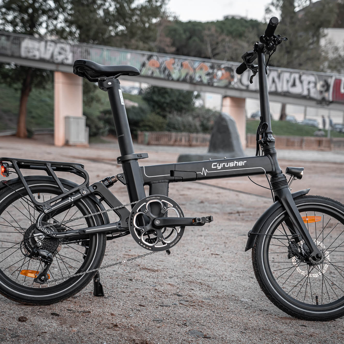 Czarny składany rower elektryczny zaparkowany na żwirze. W tle most z graffiti. Rower ma logo Cyrusher.