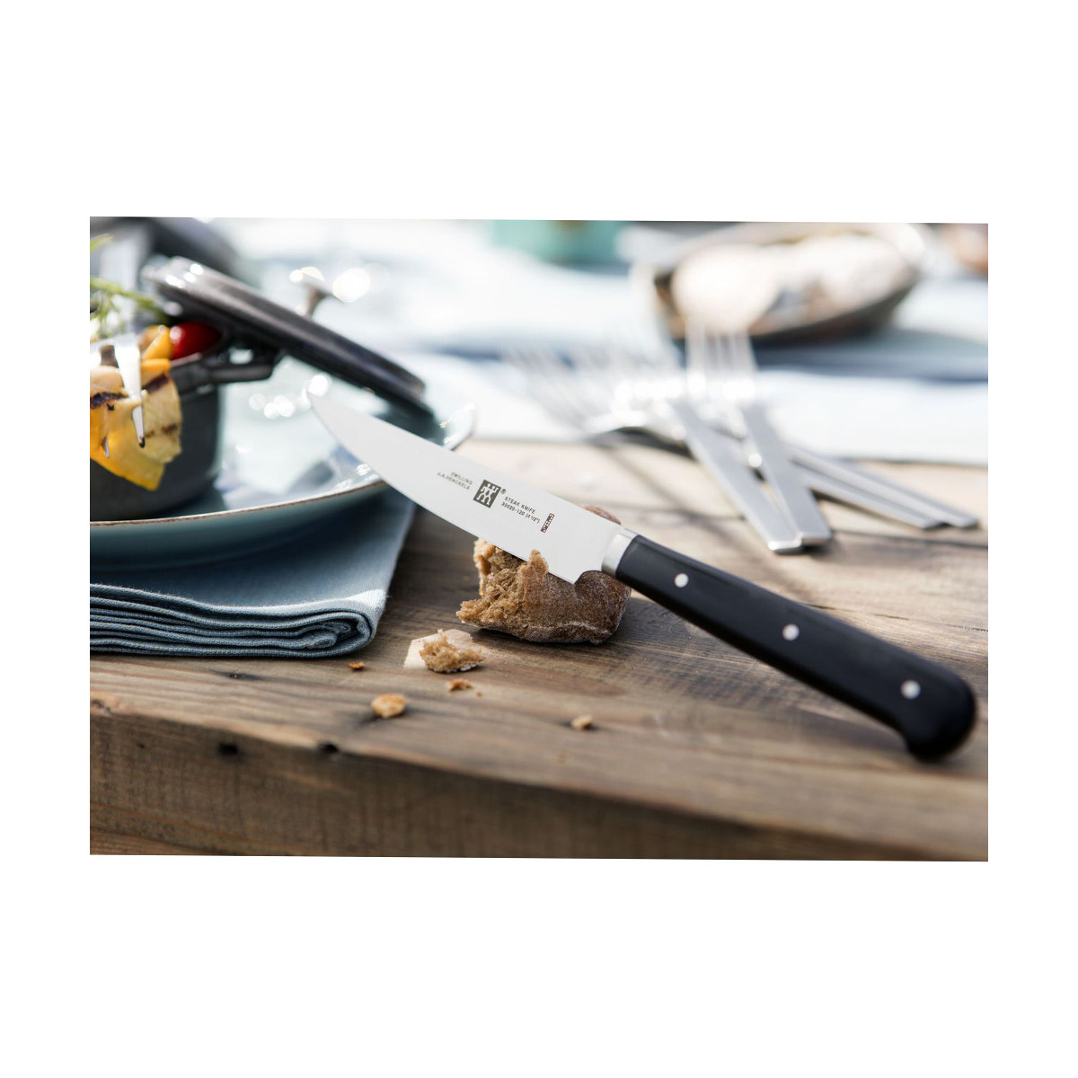 Nóż z czarną rączką leży na drewnianym stole obok okruchów chleba i kawałka chleba. W tle inne sztućce i talerze.