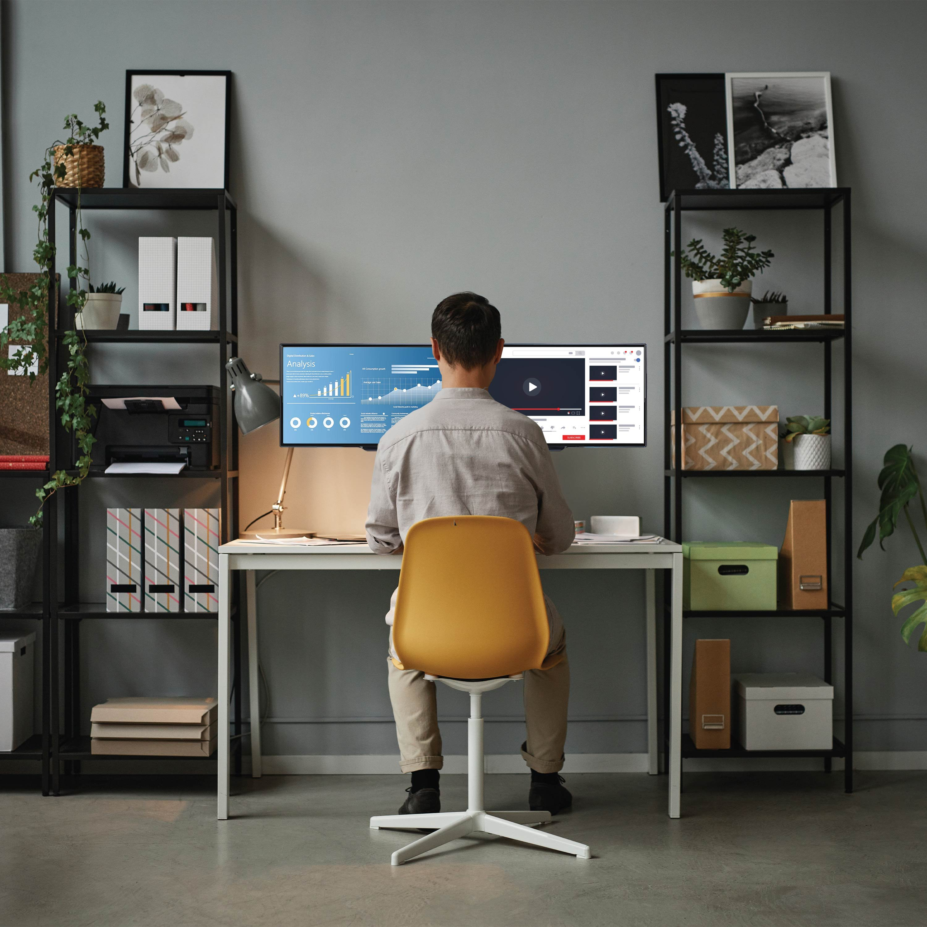 Une personne travaille sur un ordinateur à un bureau. Des étagères de chaque côté avec plantes, dossiers et décoration.