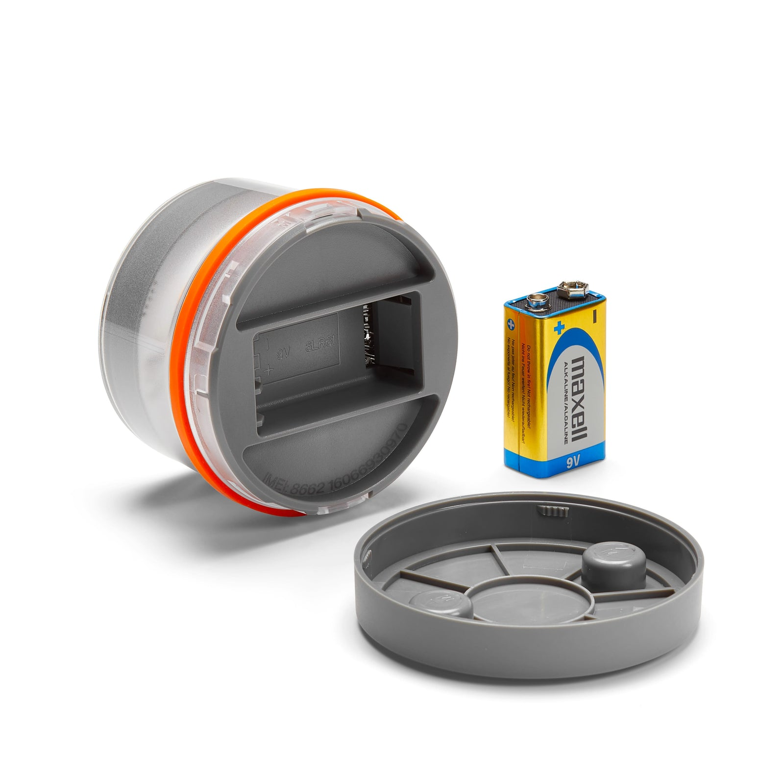 Un contenedor gris abierto con un anillo naranja, una tapa separada y una batería Maxell de 9V sobre fondo blanco.
