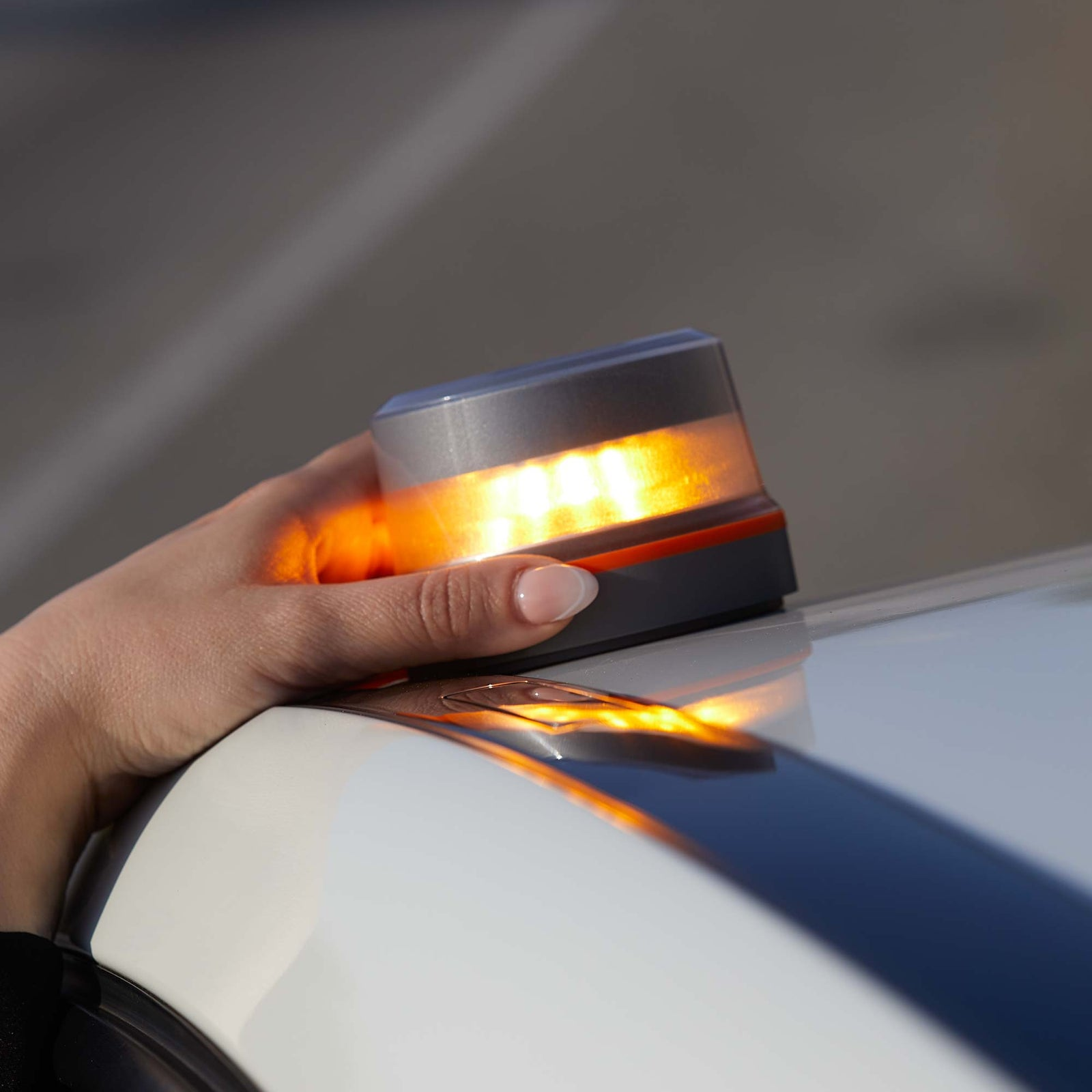 Mano sosteniendo una luz naranja intermitente en un coche blanco. La luz es cilíndrica, con tapa gris y base negra.