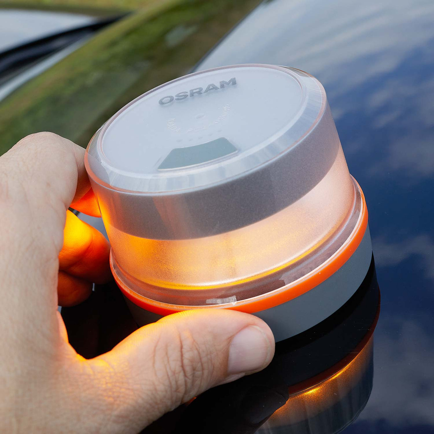 Una mano sostiene una luz de emergencia Osram naranja y gris, que se refleja en una superficie oscura y brillante. La luz está encendida.