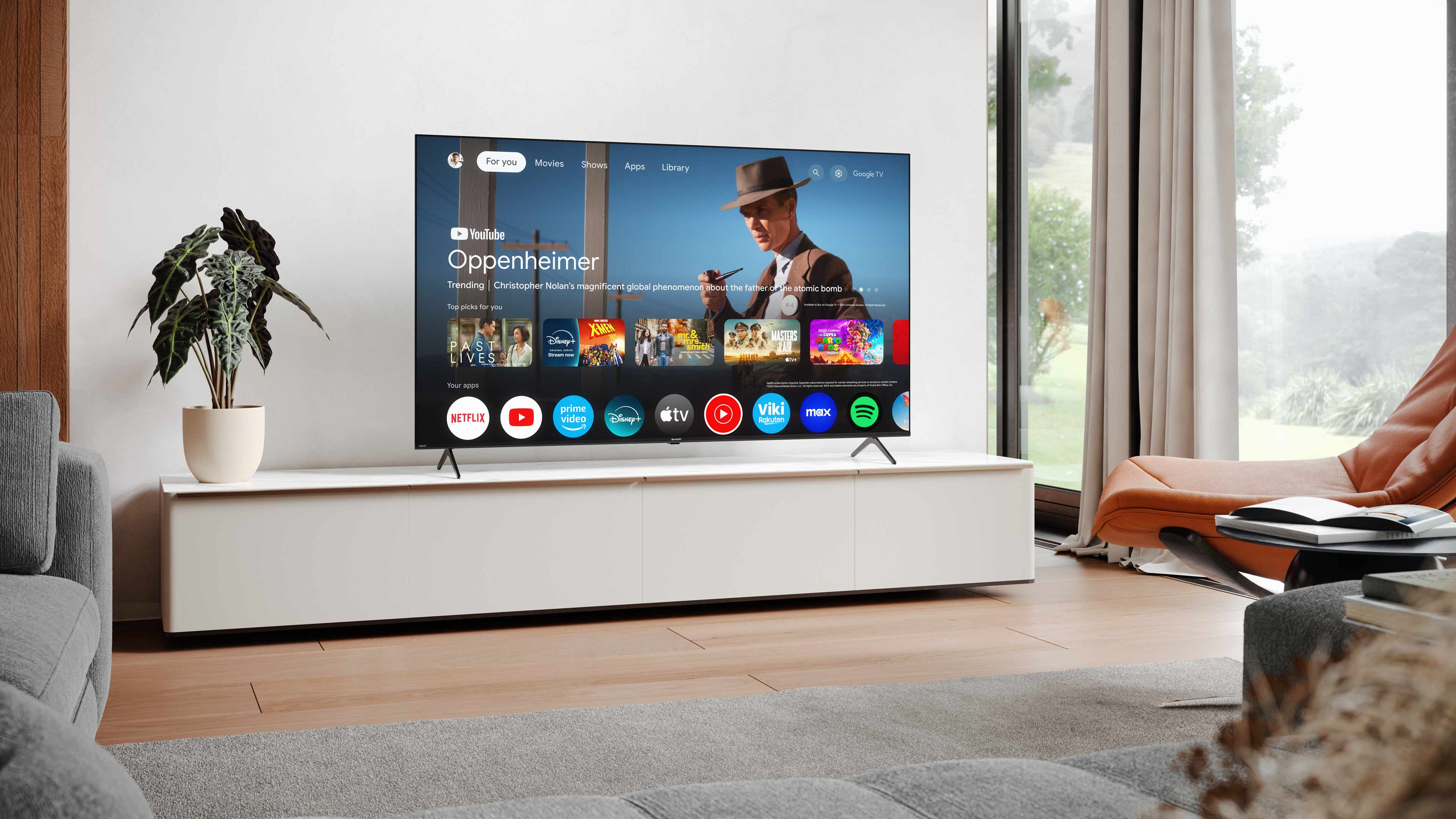 Ein modernes Wohnzimmer mit einem Fernseher, der Oppenheimer zeigt. Eine Pflanze, ein Sofa und ein Stuhl sind vorhanden. Das Zimmer hat eine weiße Wand.