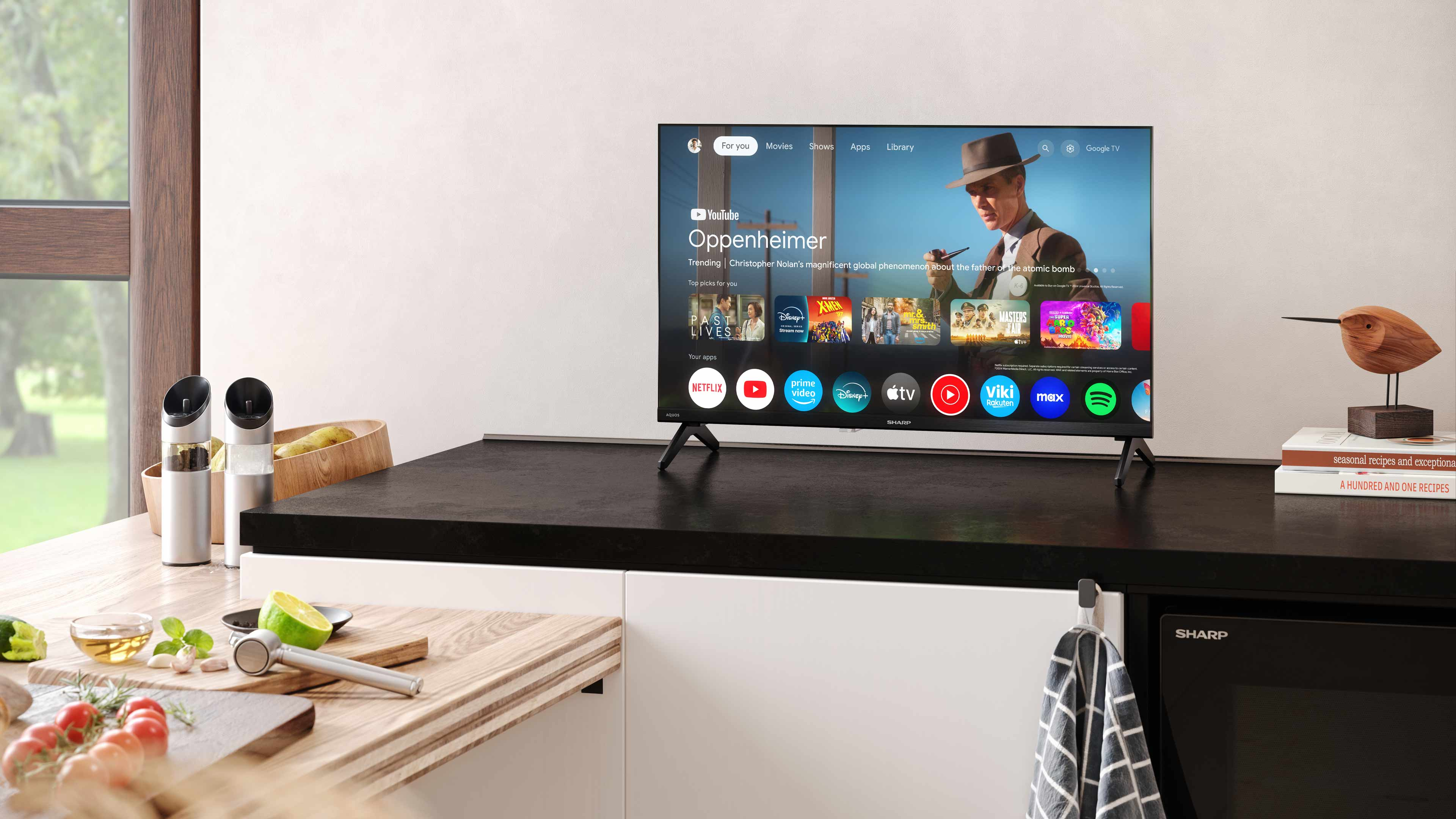 Küchenszene mit TV-Gerät, das Oppenheimer zeigt, Lebensmitteln und Küchenutensilien. Rechts eine Holzvogelstatue.