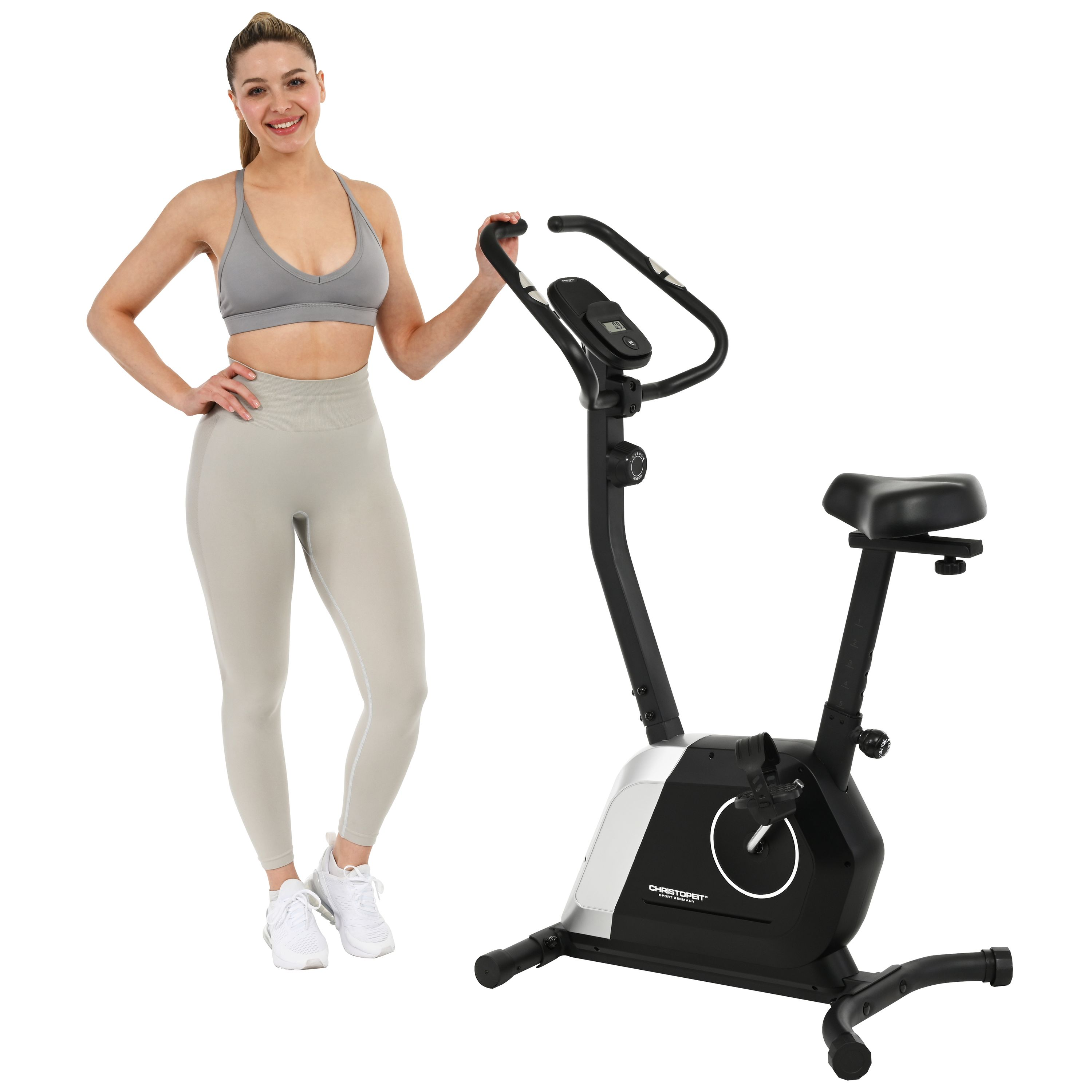 Frau in Sportkleidung posiert an einem Heimtrainer. Fahrrad ist schwarz und silber. Weißer Hintergrund.