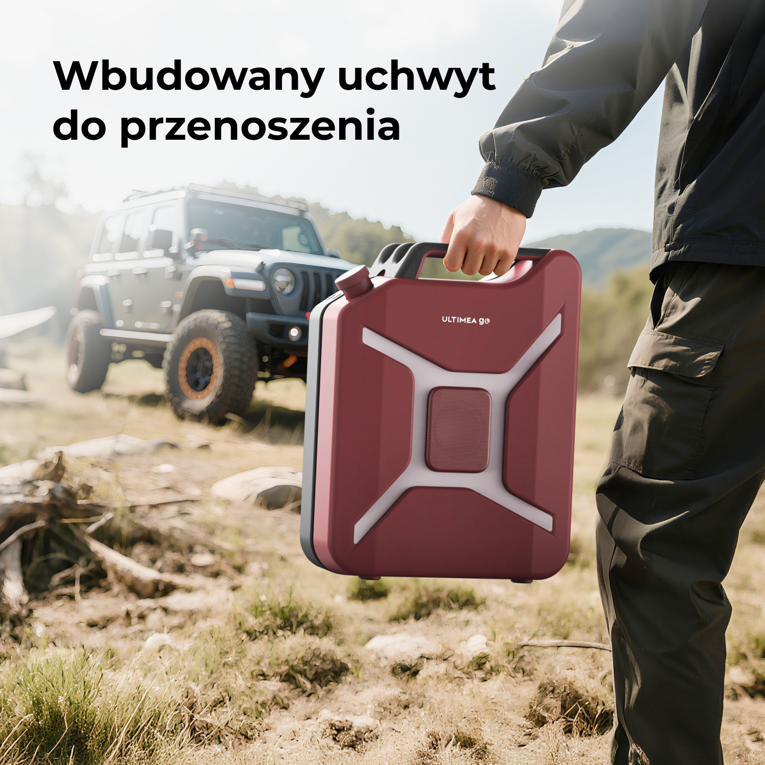 Osoba trzyma w dłoni czerwony głośnik przenośny z uchwytem. W tle jeep w naturalnym otoczeniu.