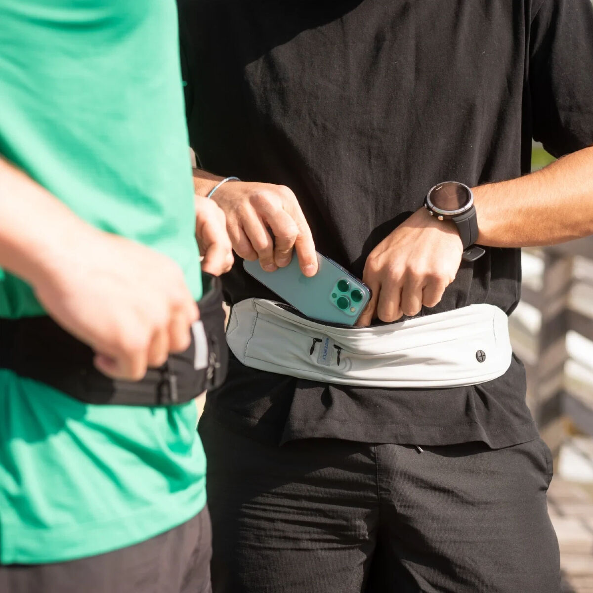 Sono mostrate due persone. Uno tiene un telefono e lo sta mettendo in una cintura bianca. L'altro indossa una camicia verde e una cintura nera.