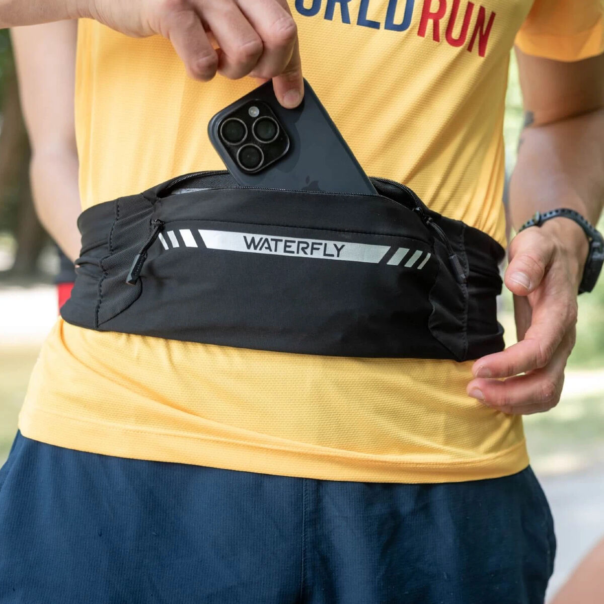Una persona sta mettendo uno smartphone in un marsupio nero. La persona indossa una maglia gialla e pantaloncini blu. La borsa ha la scritta 'WATERFLY'.