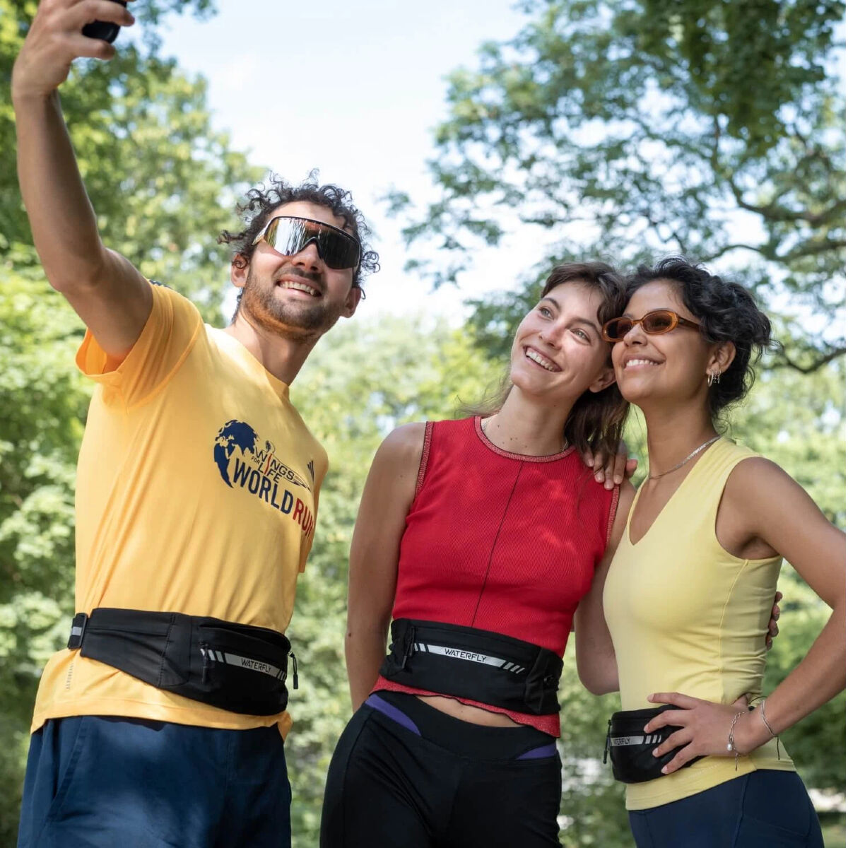 Tre persone che fanno un selfie all'aperto. Indossano occhiali da sole, abbigliamento da corsa e marsupi. Alberi sullo sfondo.
