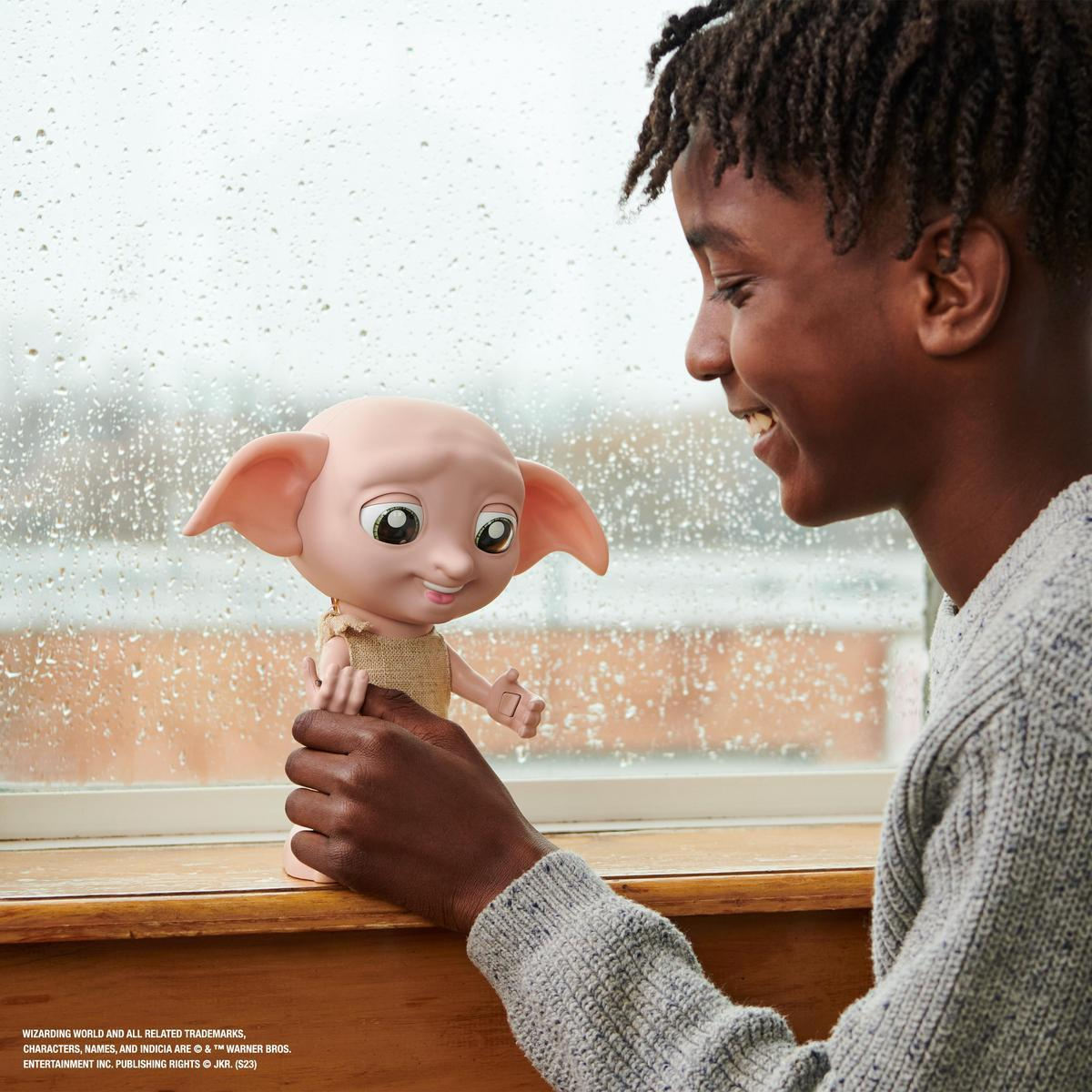 Un niño negro sonriente sostiene una muñeca de Dobby, mirándola con alegría. La muñeca tiene orejas grandes y ojos grandes. Un ventana con gotas de lluvia en el fondo.