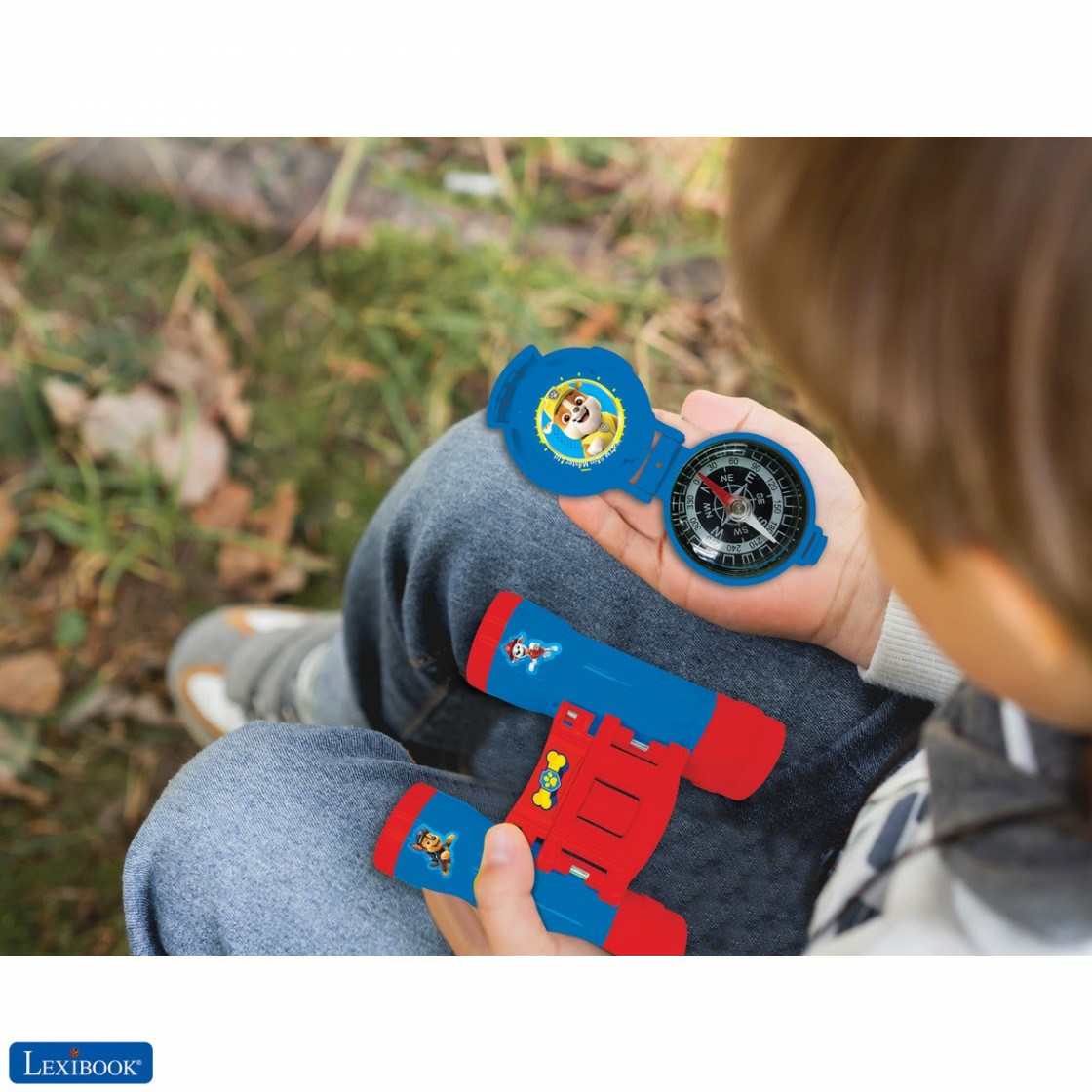 Ein Kind hält einen blauen Paw Patrol-Kompass und ein rotes Fernglas. Der Hintergrund ist eine verschwommene grün-braune Natur.