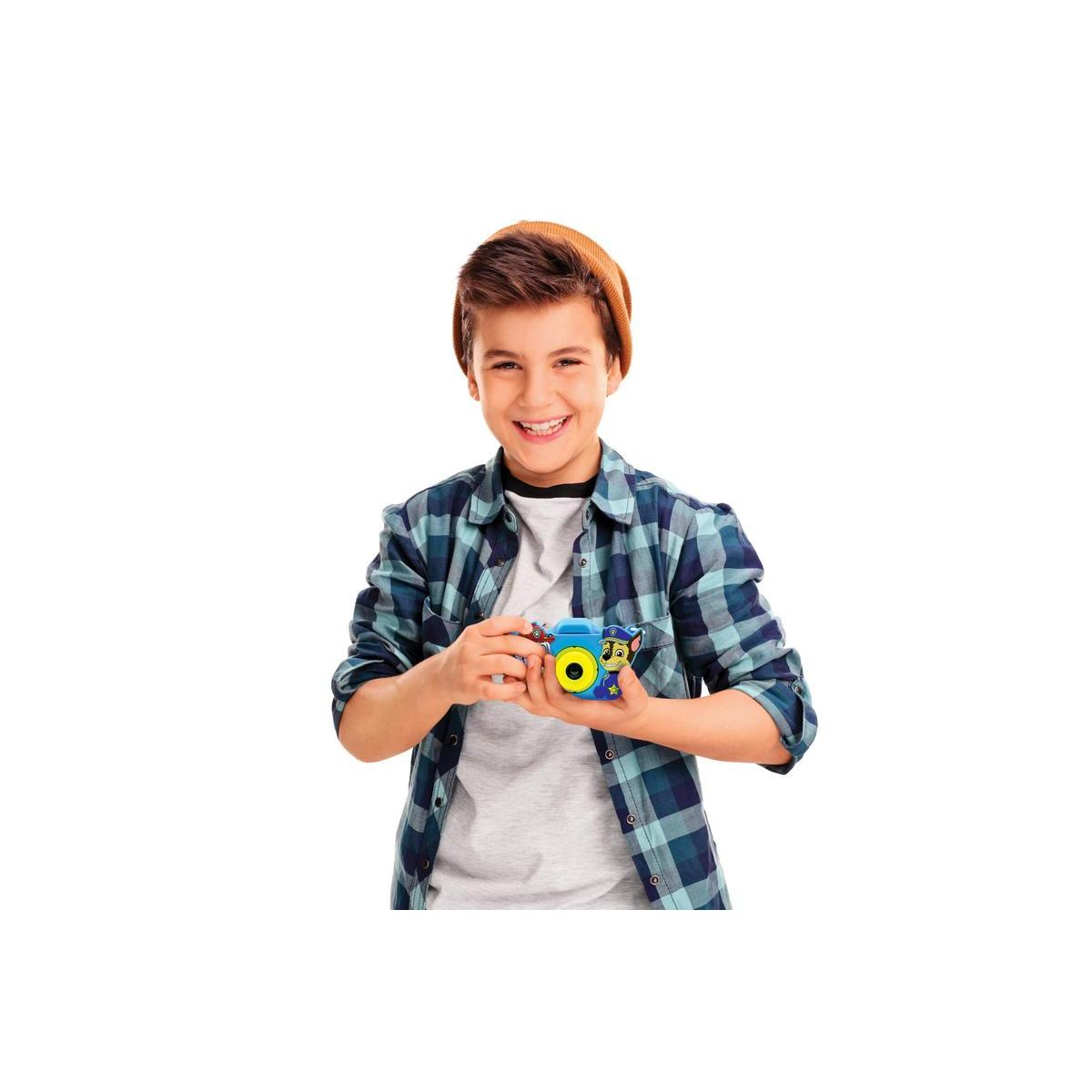 Un chico sonriente con camisa a cuadros y gorro sostiene una cámara de juguete azul con detalles amarillos.