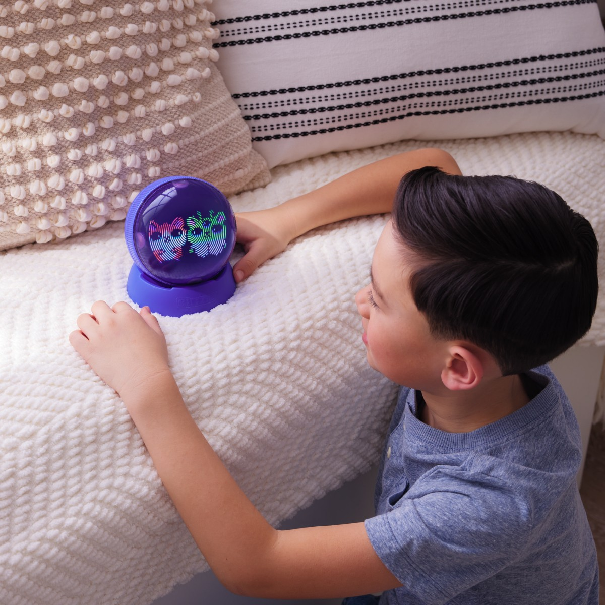 Un ragazzo guarda un dispositivo viola e trasparente illuminato su una coperta bianca. I cuscini sono sullo sfondo. Il ragazzo indossa una camicia blu.