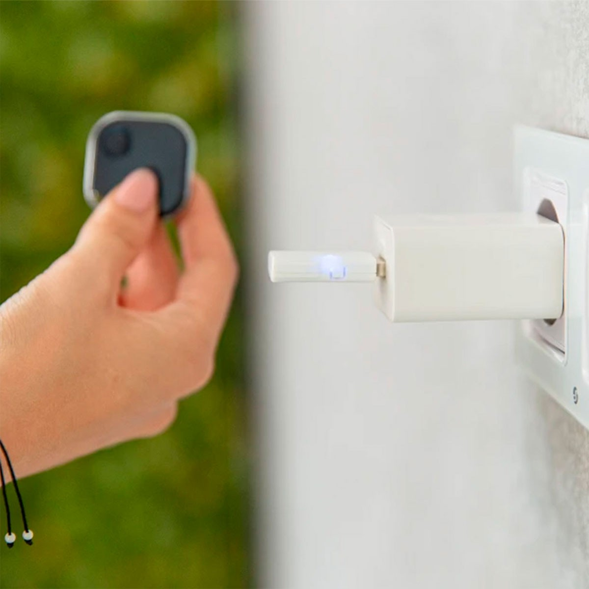 Une main tenant un appareil carré noir près d'un chargeur blanc branché dans une prise murale. Arrière-plan vert flou.
