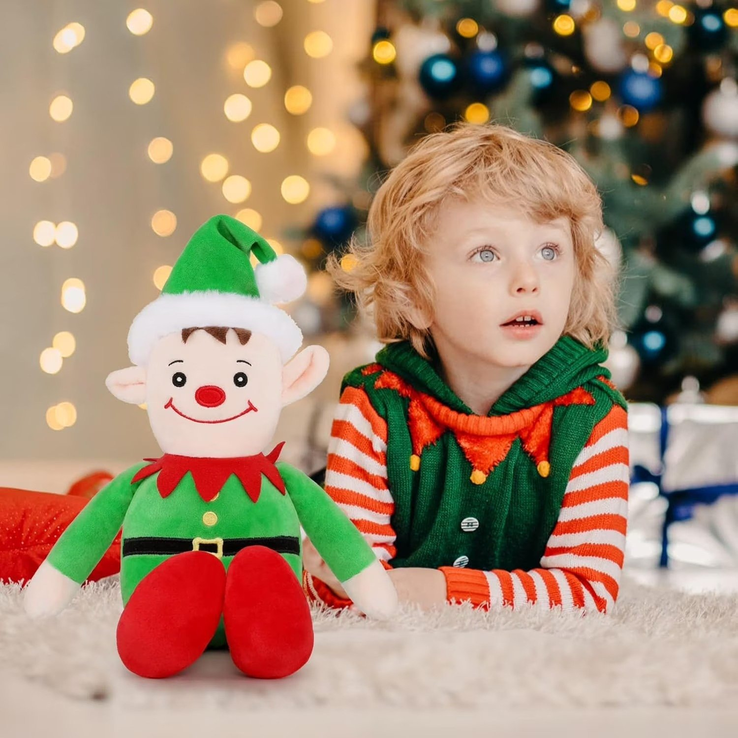 Un bambino e una bambola elfo vicino a un albero di Natale. Il bambino indossa un costume da elfo. La bambola ha un cappello verde e scarpe rosse.