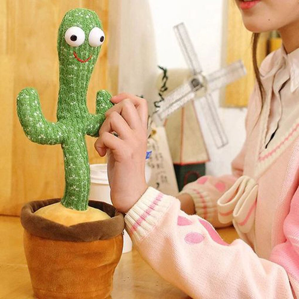 Una persona sostiene un juguete de cactus verde con ojos grandes y una boca sonriente. El juguete está en una maceta marrón, con un molino de viento de fondo.