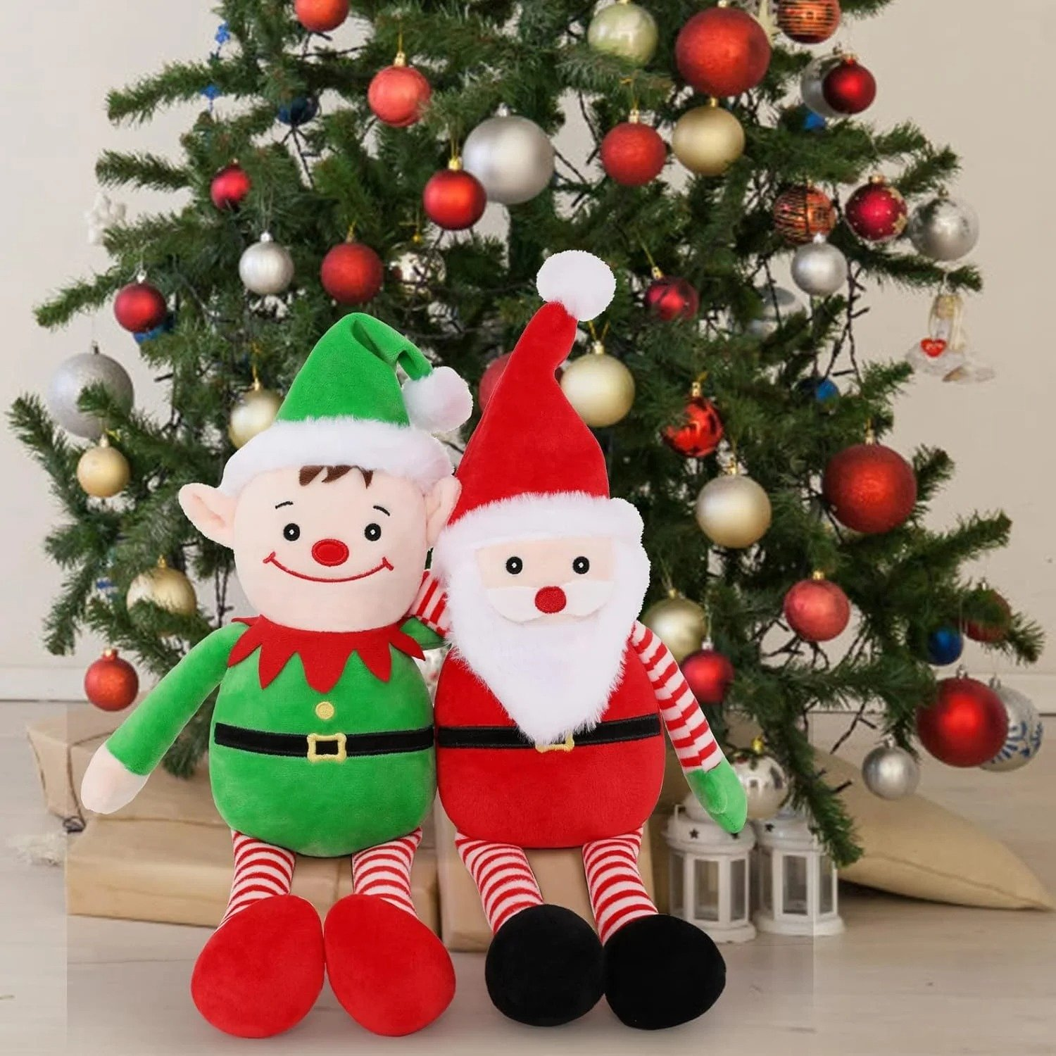 Due peluche natalizi, un elfo e Babbo Natale, sono seduti davanti a un albero decorato con ornamenti rossi e dorati.