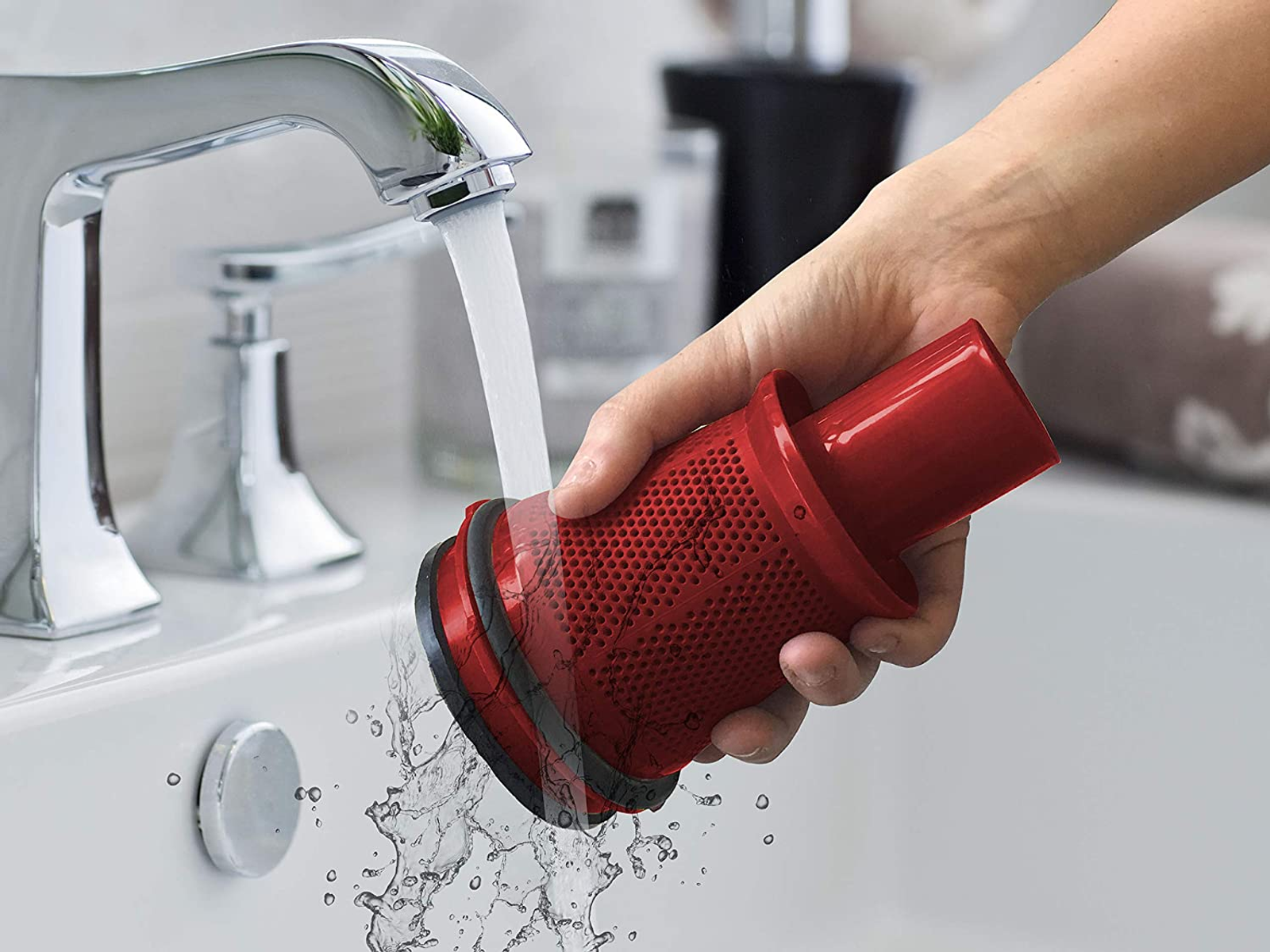 Una mano tiene un filtro rosso per aspirapolvere sotto l'acqua corrente di un rubinetto cromato in un bagno.