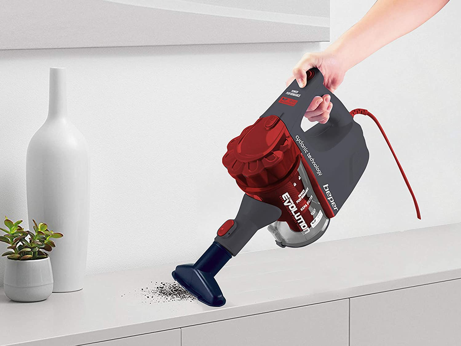 Una mano tiene un aspirapolvere rosso e grigio, pulendo una superficie bianca. Sono presenti anche un vaso bianco e una pianta.
