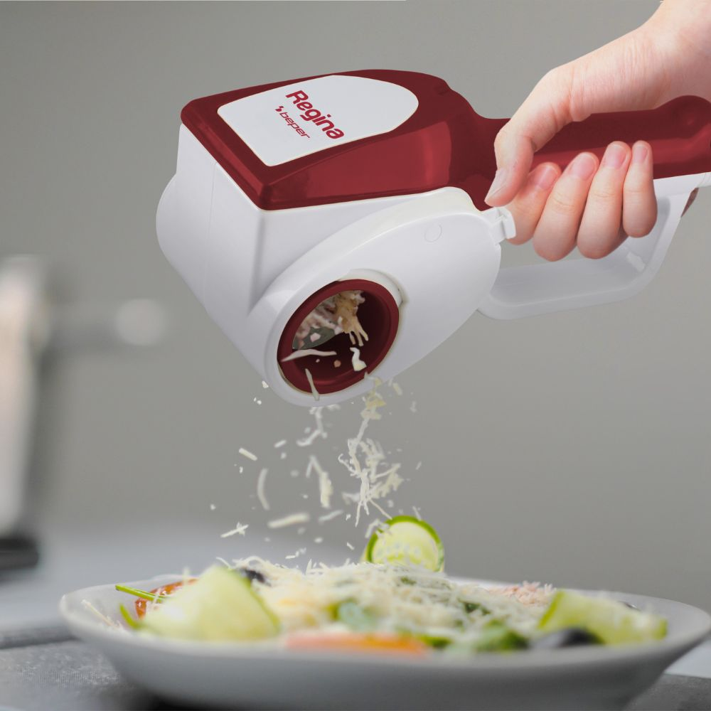 Una mano grattugia formaggio su un'insalata. La grattugia è rossa e bianca, con l'etichetta 'Regina'. Il formaggio cade su un piatto di insalata.
