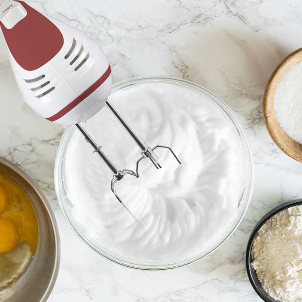Un frullatore a mano bianco e rosso sta mescolando albumi in una ciotola di vetro. Ci sono ciotole di zucchero, farina e uova nelle vicinanze.