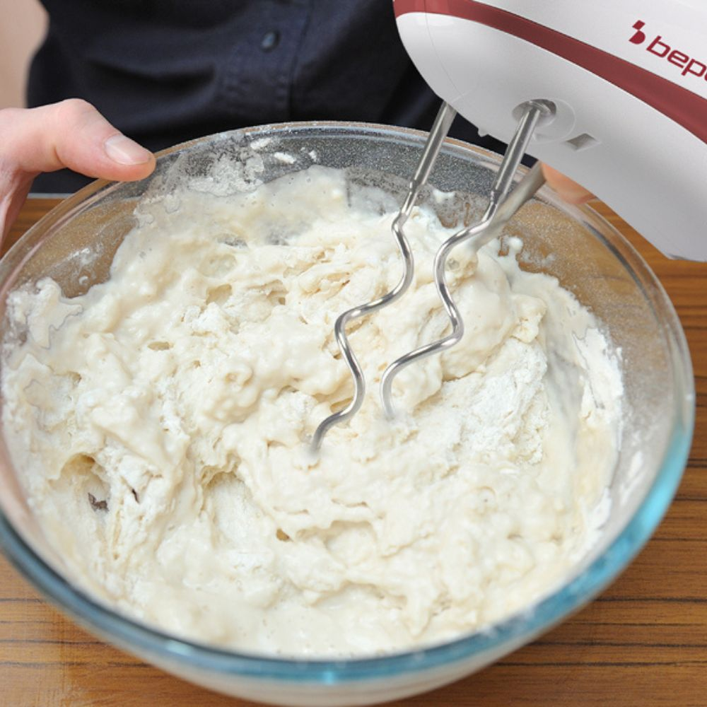 Un frullatore a mano sta mescolando l'impasto in una ciotola di vetro. L'impasto è bianco. Una mano tiene la ciotola.