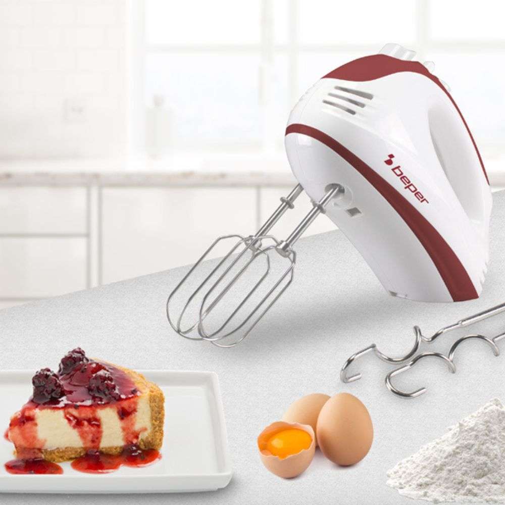 Un frullatore a mano bianco e rosso, cheesecake, uova e farina su un tavolo. Il frullatore è di Beper. Una fetta di cheesecake con topping di bacche.