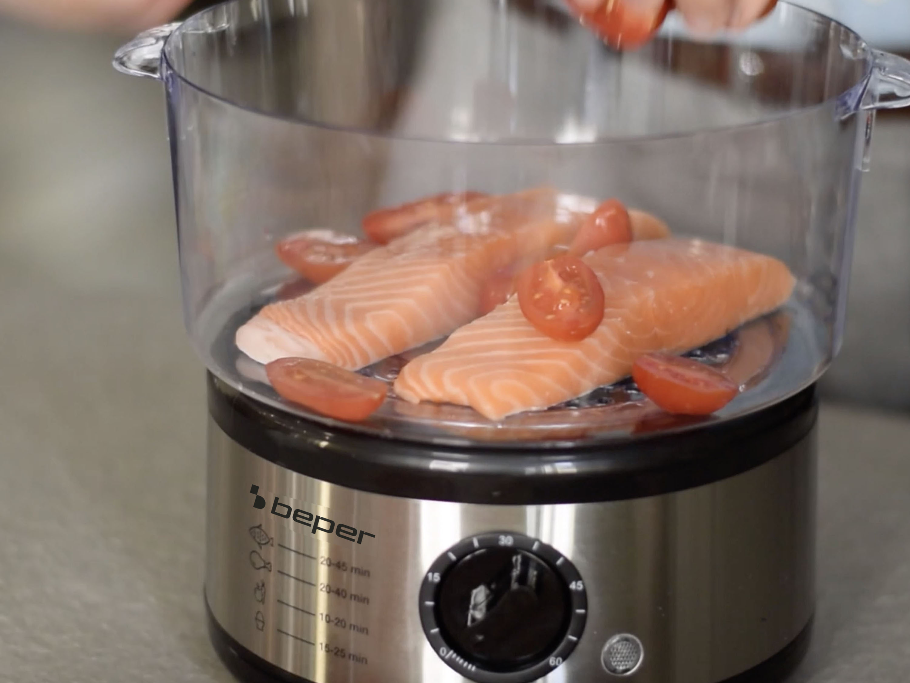 Filetti di salmone e pomodorini in vaporiera. Una mano aggiunge altri pomodori. Base in acciaio inox.