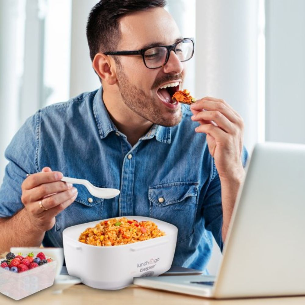 Un uomo con gli occhiali mangia cibo da un contenitore su una scrivania con un laptop. Sono presenti anche un portapranzo bianco e un contenitore di frutta.