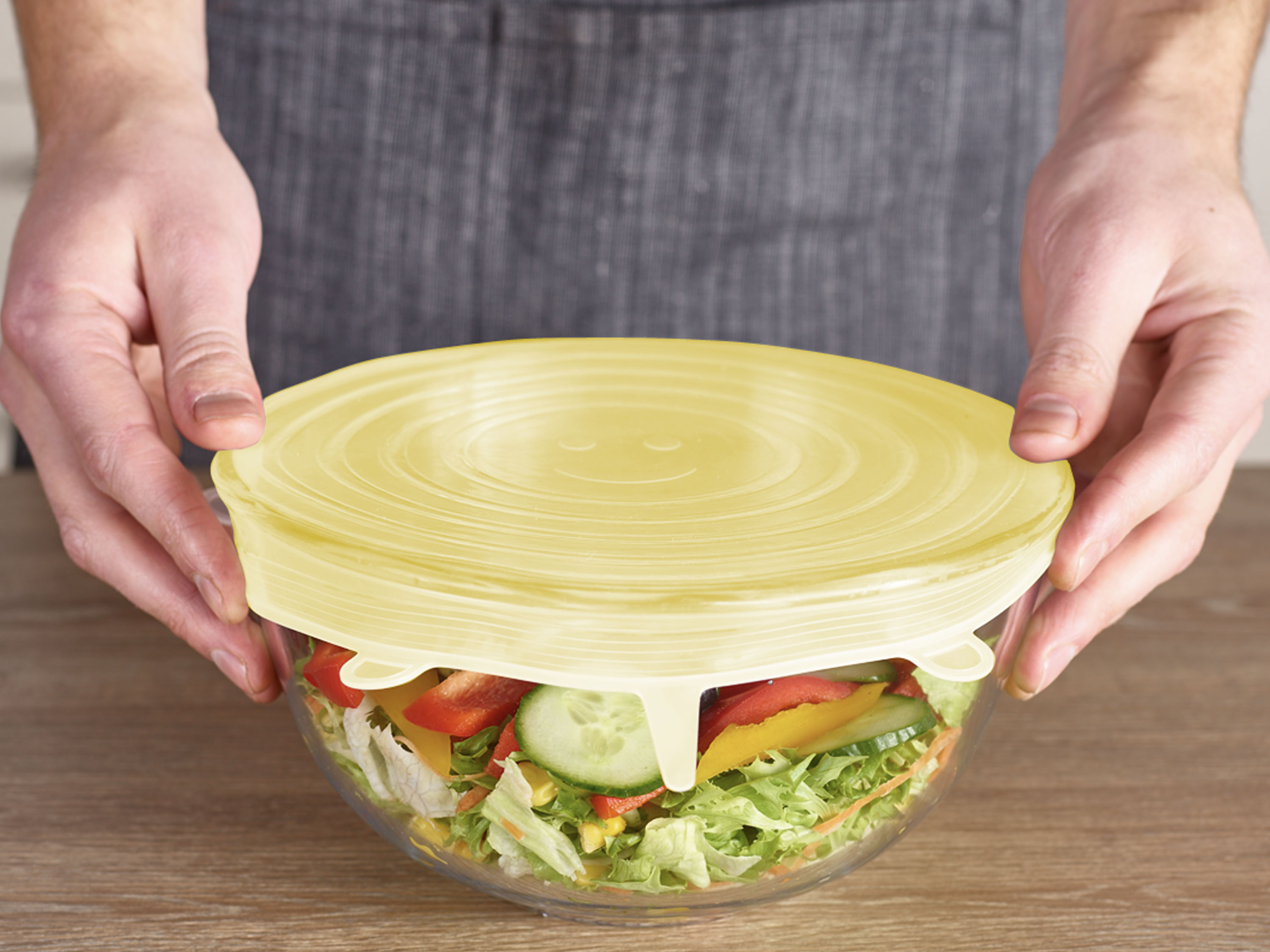 Mani che coprono una ciotola di insalata con coperchio giallo. L'insalata contiene lattuga, cetrioli, peperoni rossi e gialli.