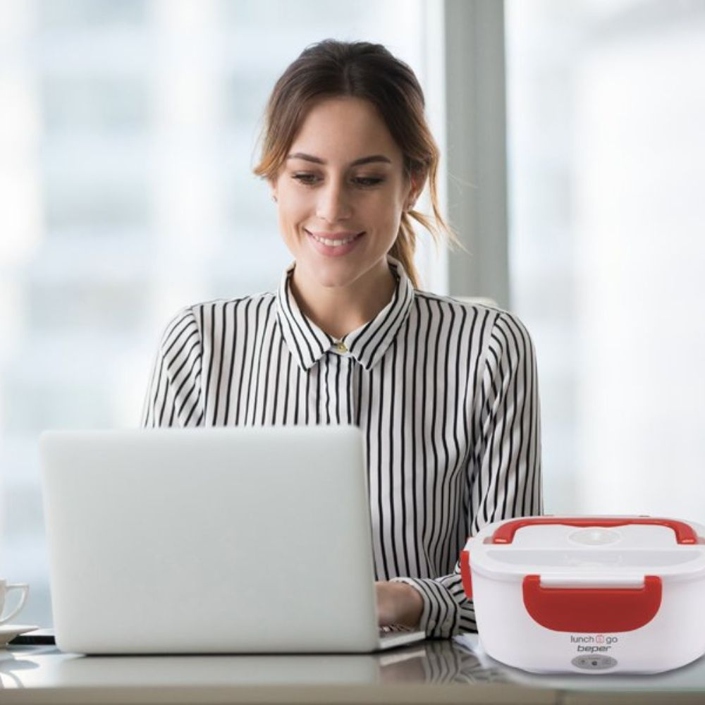 Una donna sorride mentre usa un laptop. Un portapranzo bianco con manico rosso è sul tavolo. Indossa una camicia a righe.