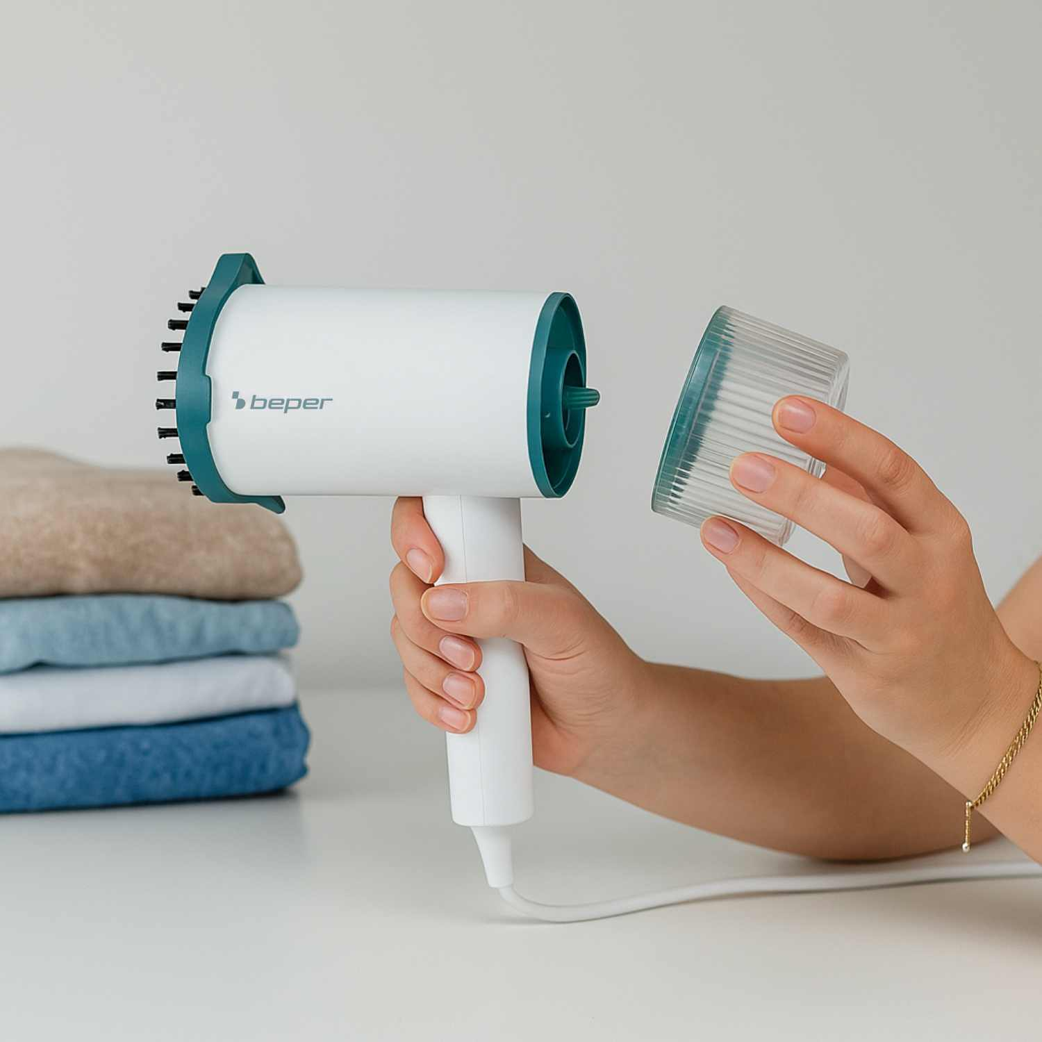 Una persona tiene in mano uno steamer per vestiti bianco e verde acqua e una parte in plastica trasparente. A sinistra c'è una pila di vestiti piegati.