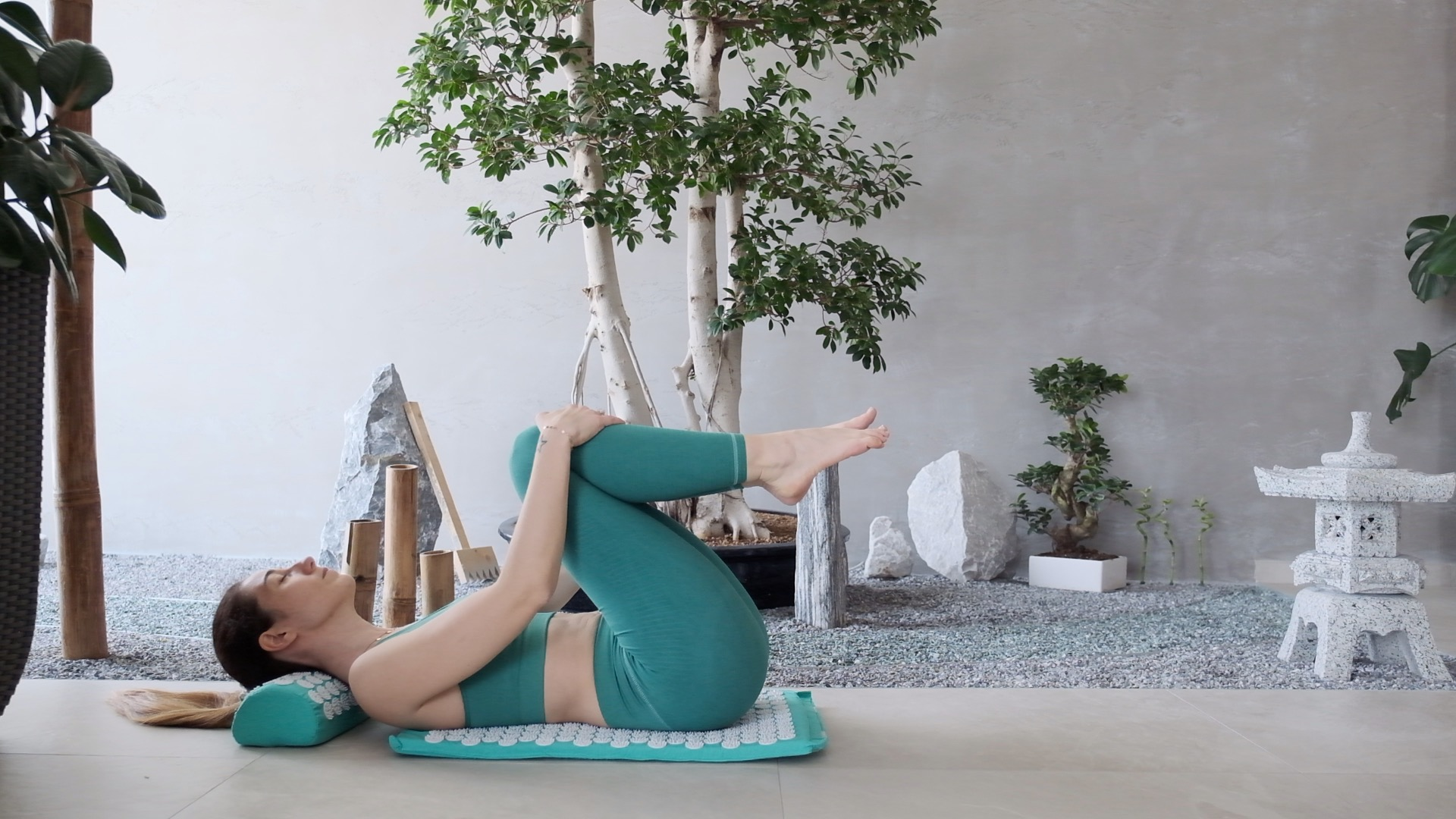 Una donna sdraiata su un tappetino, tiene le ginocchia. Indossa abbigliamento sportivo verde acqua. Un albero e elementi decorativi sullo sfondo.