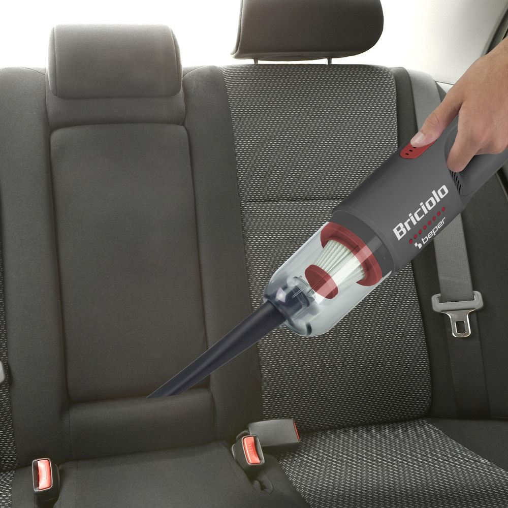 Un aspirapolvere a mano sta aspirando un sedile di un'auto. L'aspirapolvere è grigio e rosso, con il marchio 'Briciola'. Il sedile è grigio scuro.