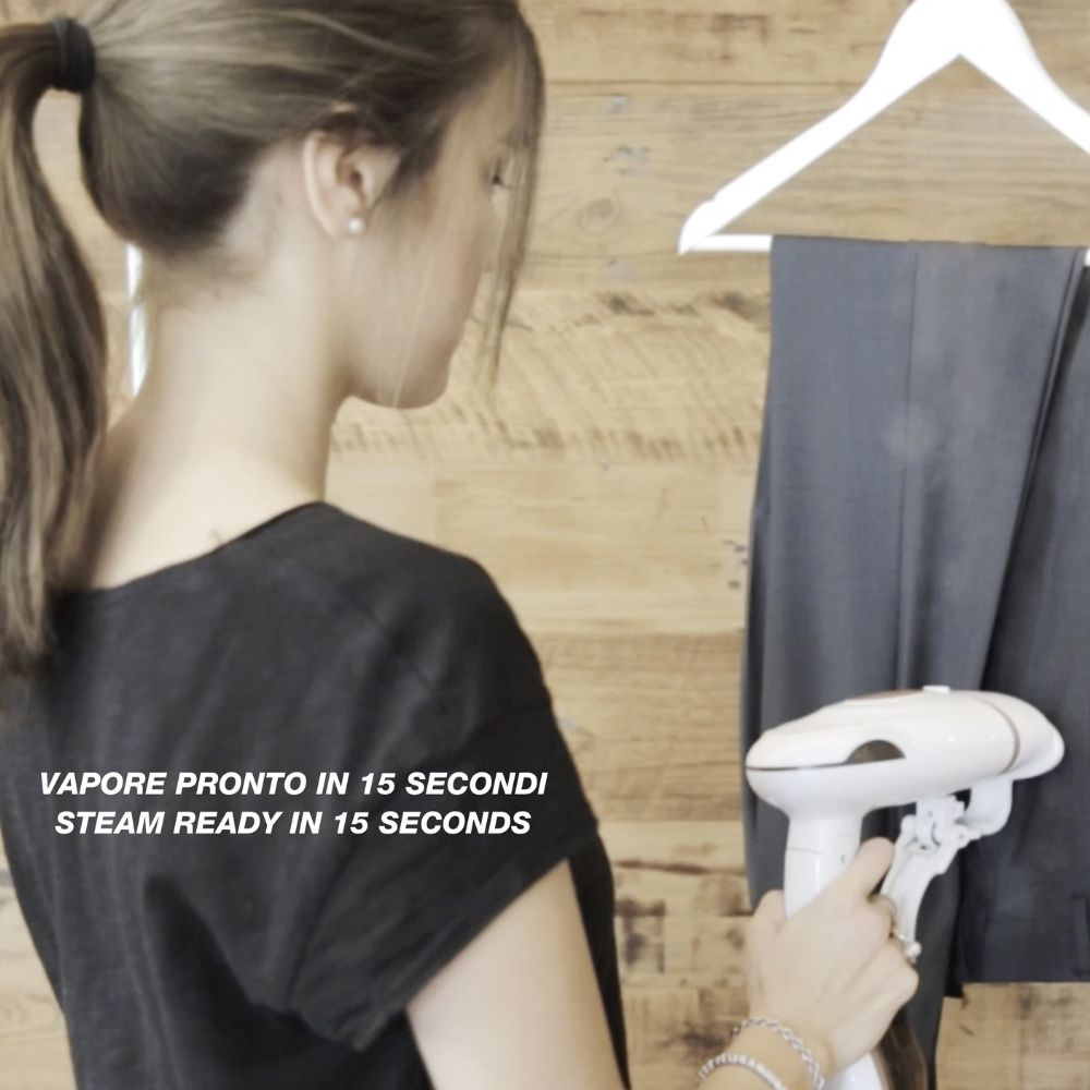 Una donna usa un vaporizzatore bianco su pantaloni grigi appesi a una parete di legno. Testo: 'Steam ready in 15 seconds'.