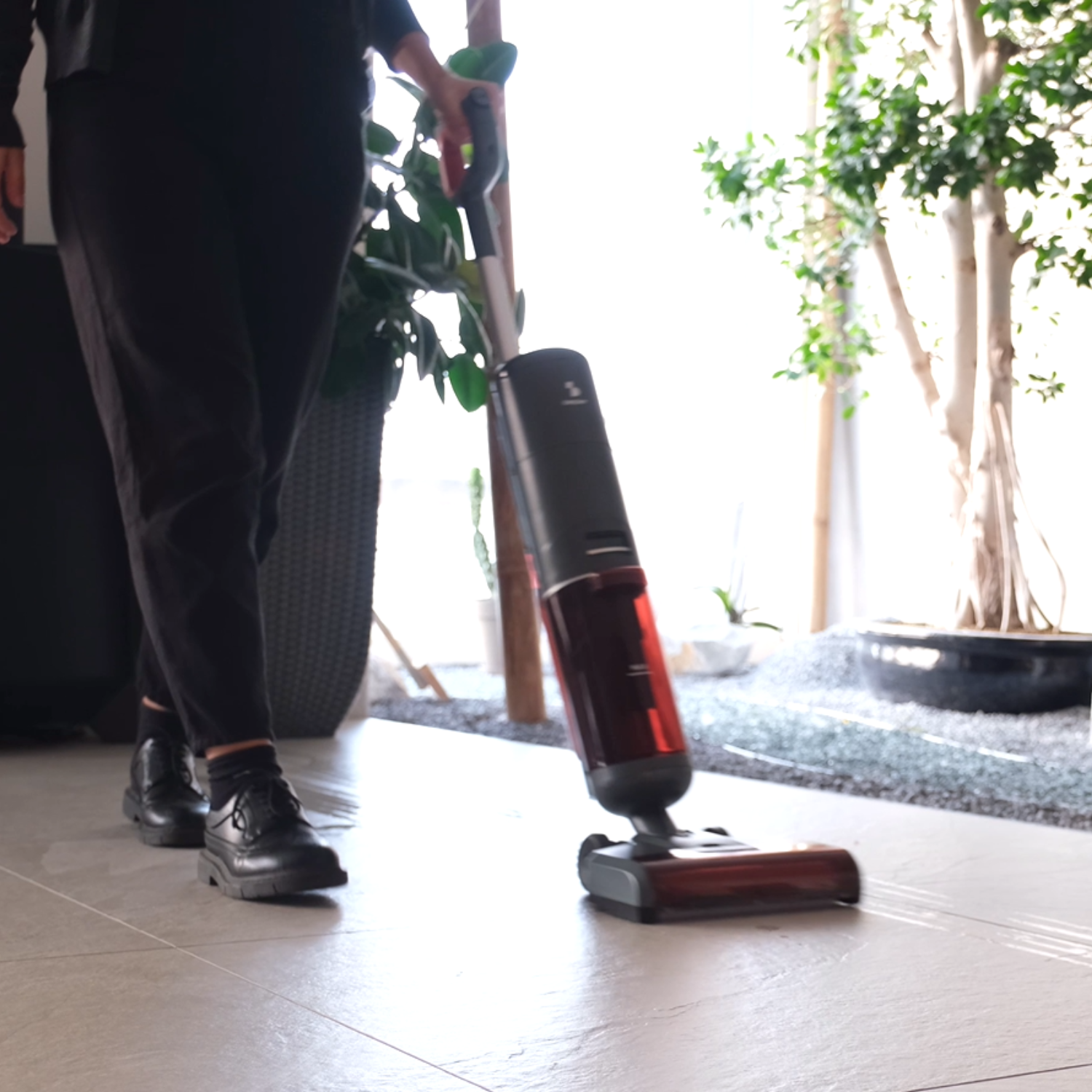 Una persona passa l'aspirapolvere. Indossa pantaloni e scarpe nere. Viene utilizzato un aspirapolvere rosso e nero. Sullo sfondo ci sono piante verdi.