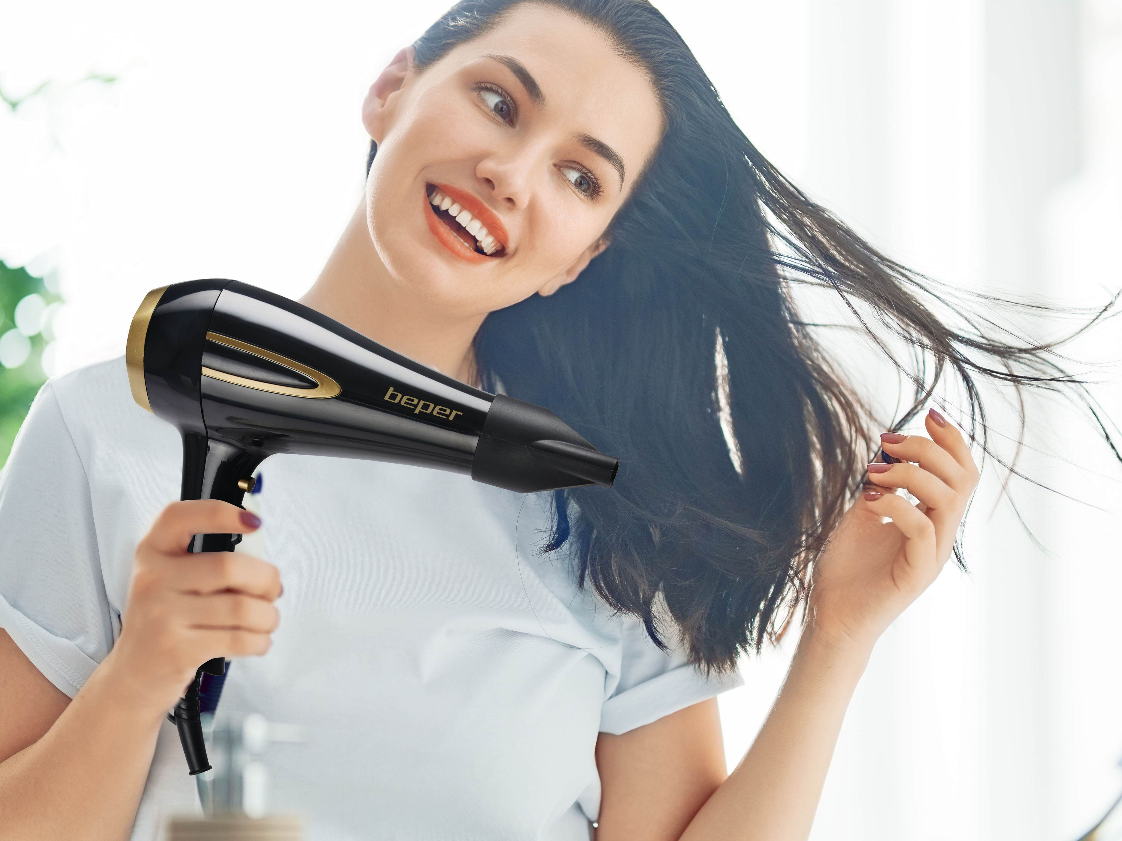 Una donna sorride, usando un asciugacapelli nero. I suoi capelli scuri fluiscono. Indossa una camicia bianca.