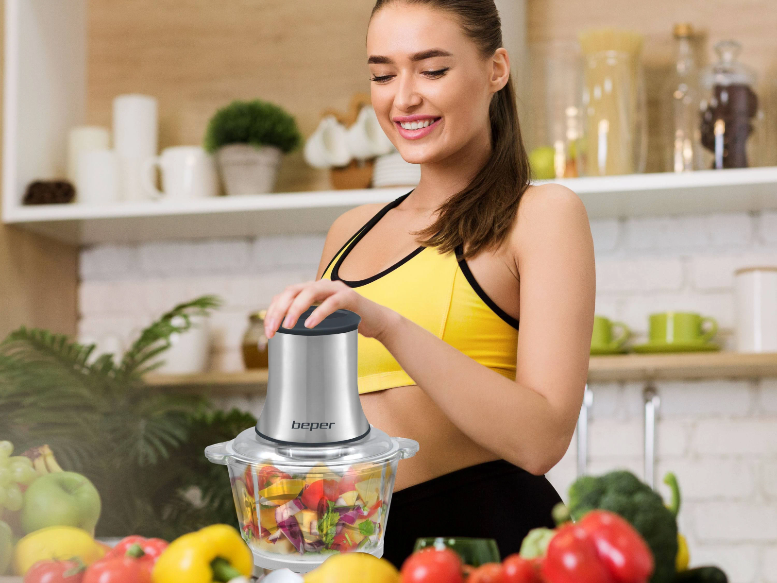 Donna in cucina con robot da cucina. Sorride, indossa un top giallo. Verdure sul bancone.