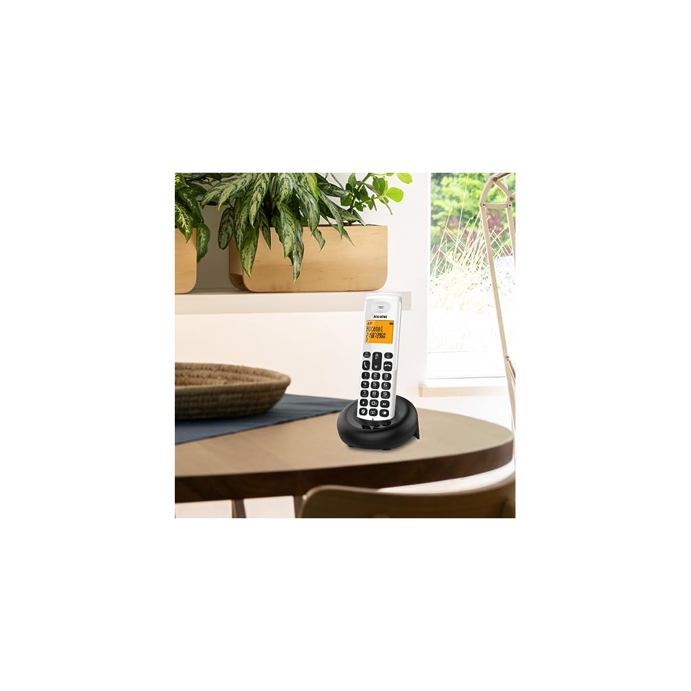 Un telefono cordless bianco su una base nera è su un tavolo di legno. Piante sullo sfondo, un cestino di vimini e una sedia visibili.